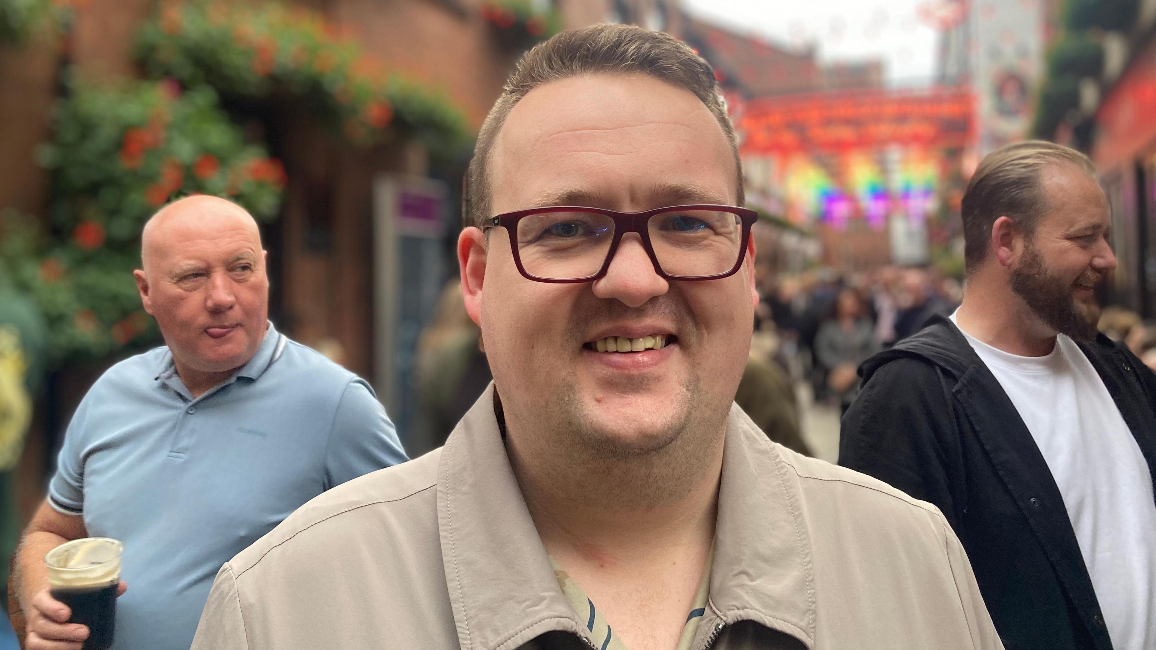 Michael Fenton, a man with short, blond hair and glasses stands smiling at the camera in a Belfast street.  He is wearing a beige jacket. Two men are standing behind him, one is holding a plastic pint of stout. 