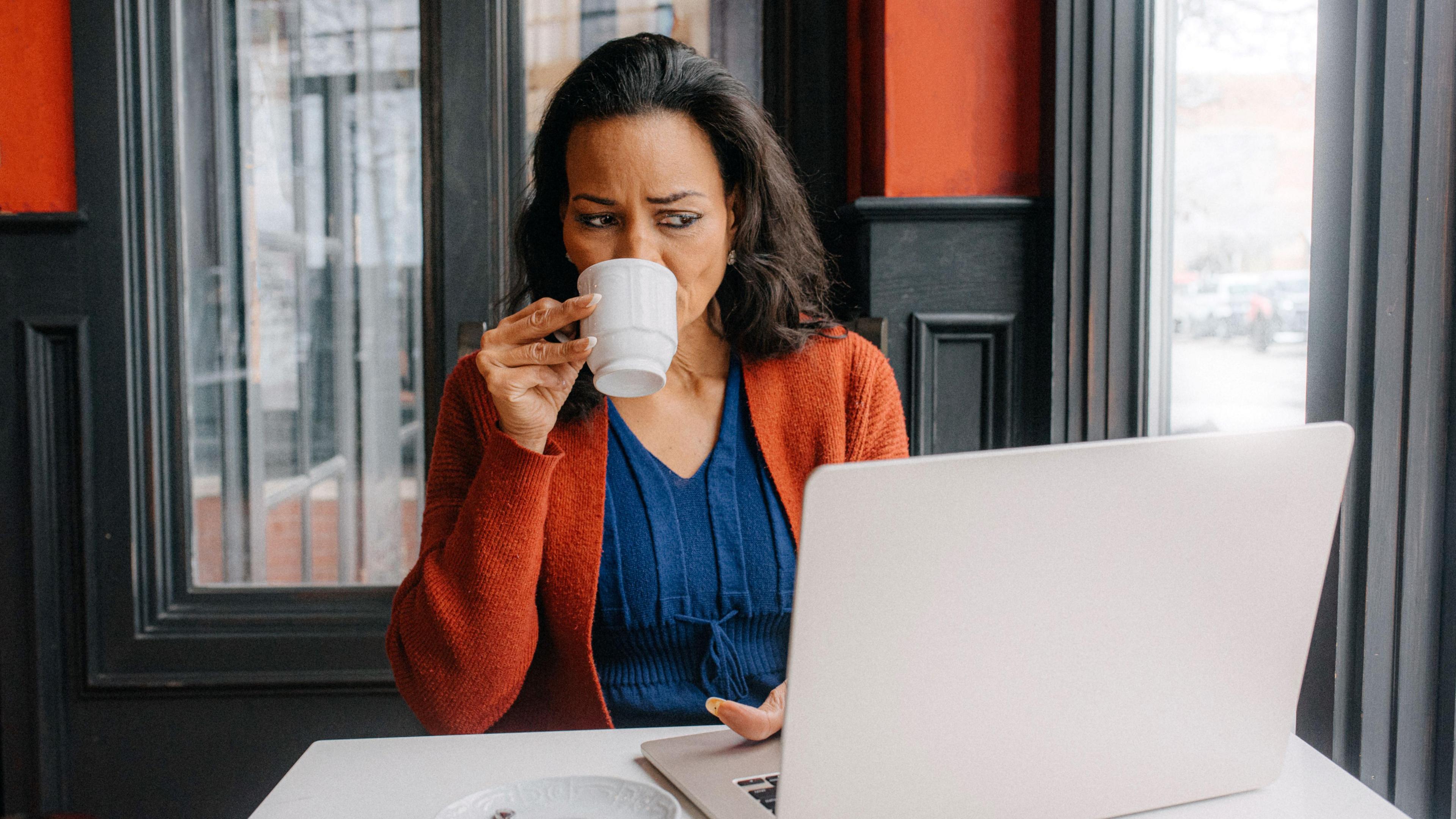 A woman is sipping from a white mug while working on her laptop at a cafe. She appears to be focused, surrounded by a warm interior with red and black accents.