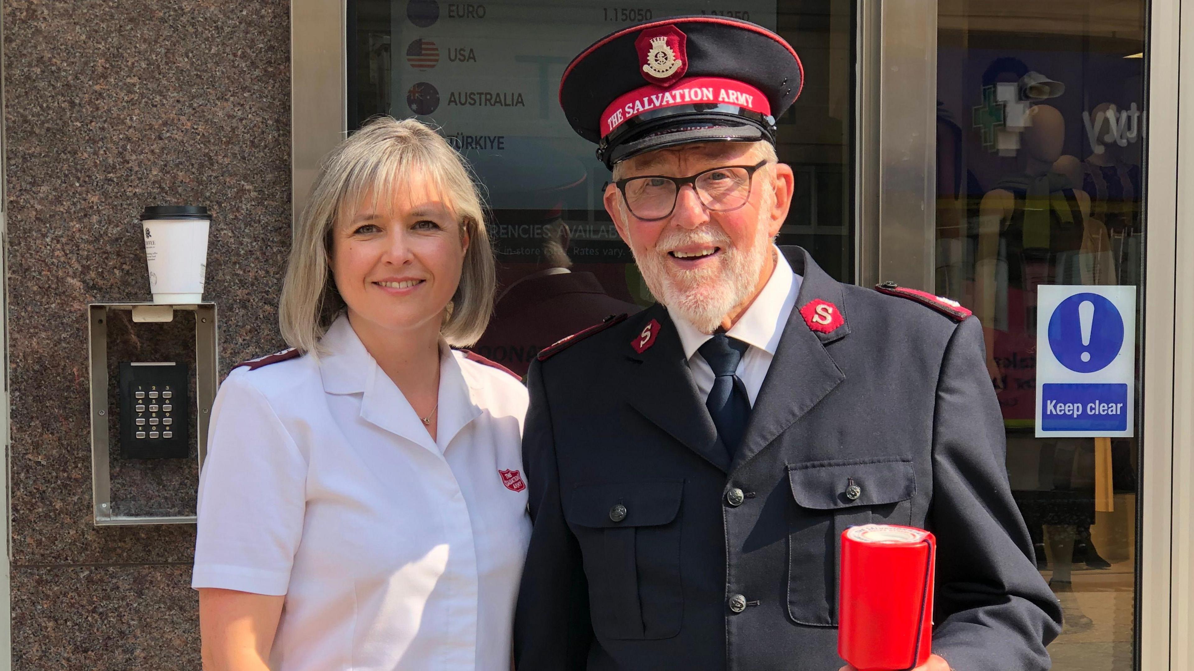 Major Leanne Cordner on the left wearing a white shirt standing next to Major John Mott who is wearing a black Salvation Army uniform with a hat and is holding a red charity collection pot. 