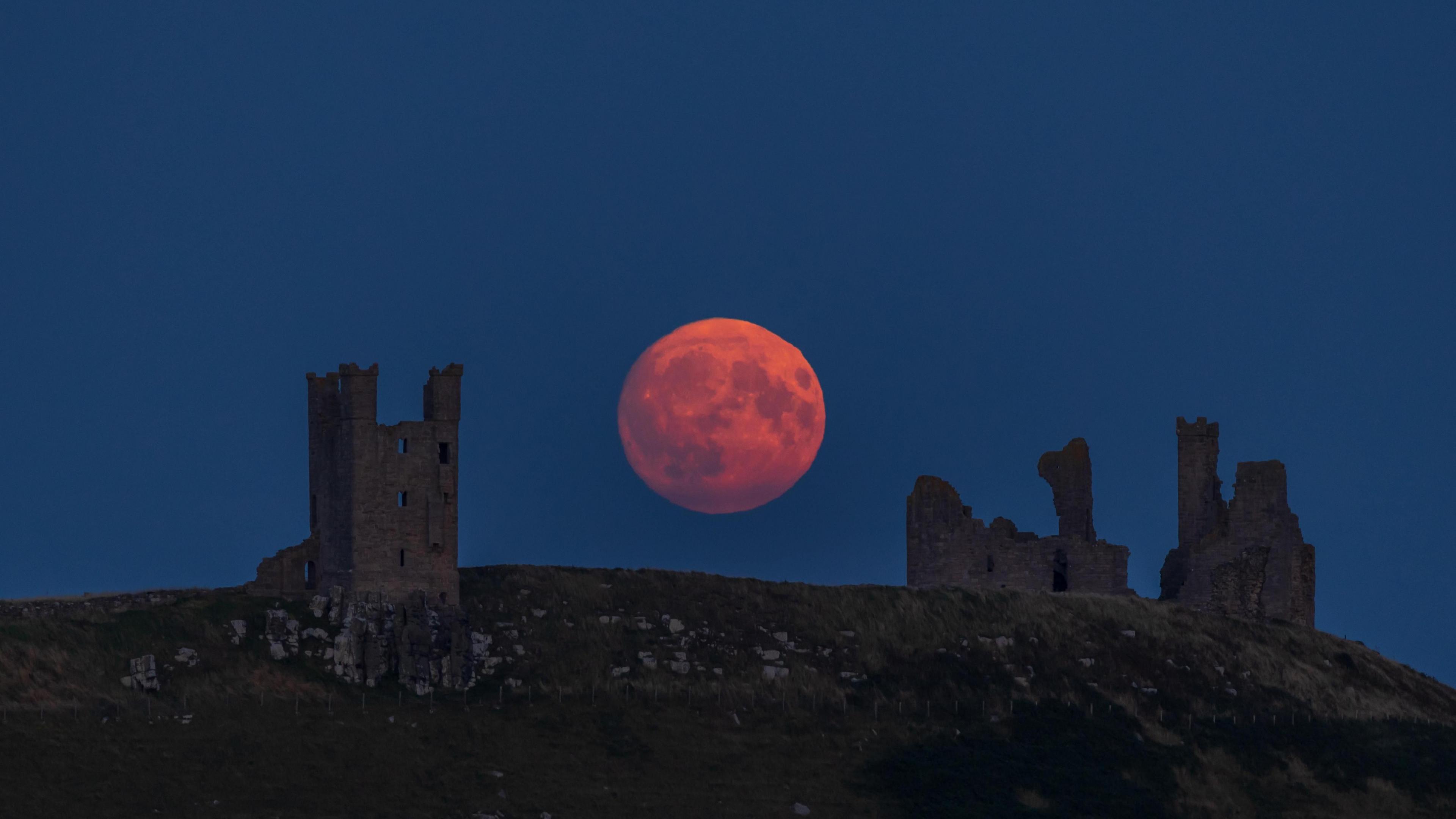 Going dark in the evening so we can just make out some castle ruins on top of a hill but behind in the background, the full moon rising low in the sky has a red tinge.