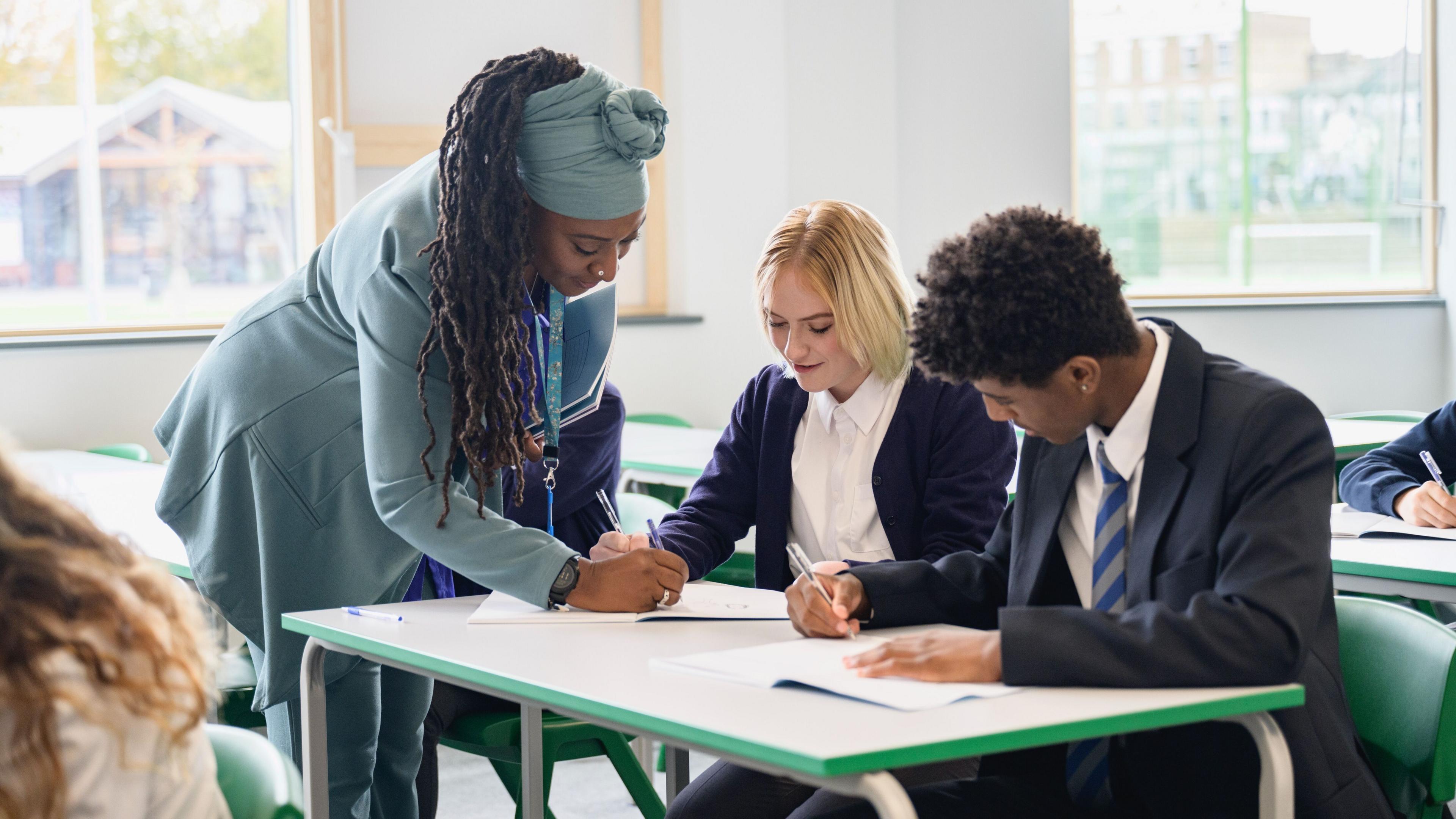 A female teacher with long braids in a pale blue coat with a matching blue headwrap, helping a blonde student in a white shirt and a male student with a white shirt and blue striped tie their school work. Sat in a white classroom with windows and green chairs.