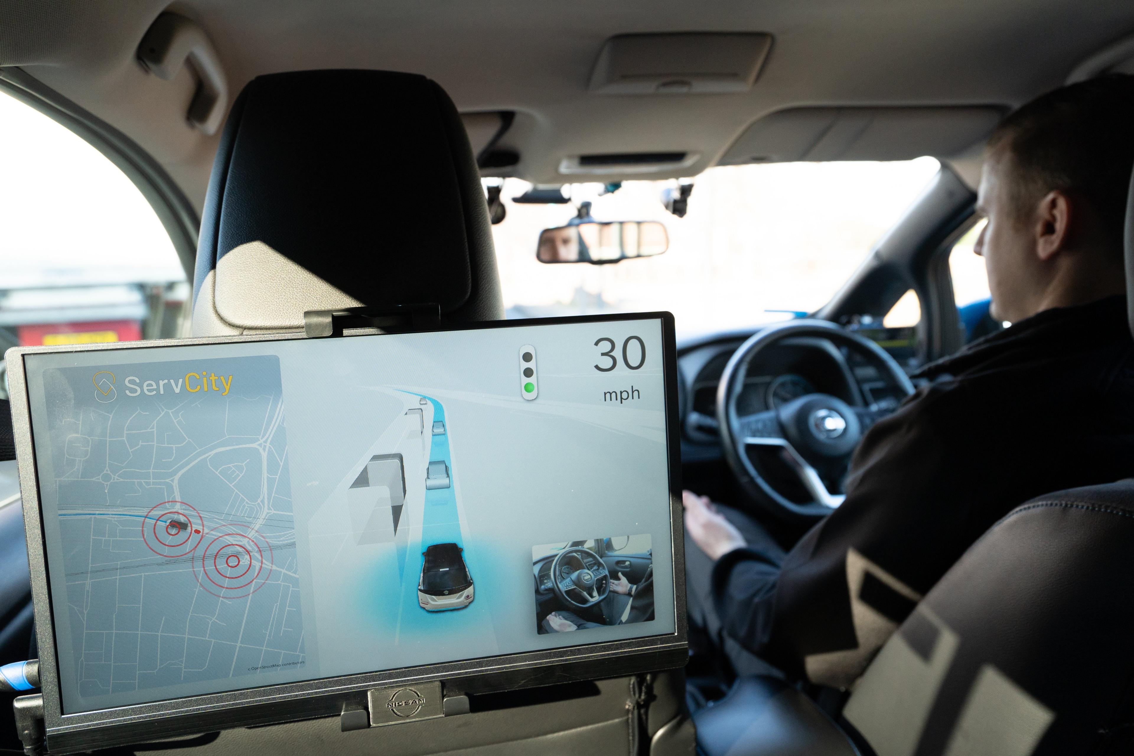 A shot inside a car with a screen showing the computer's view of the car, and a driver whose hands are off the steering wheel