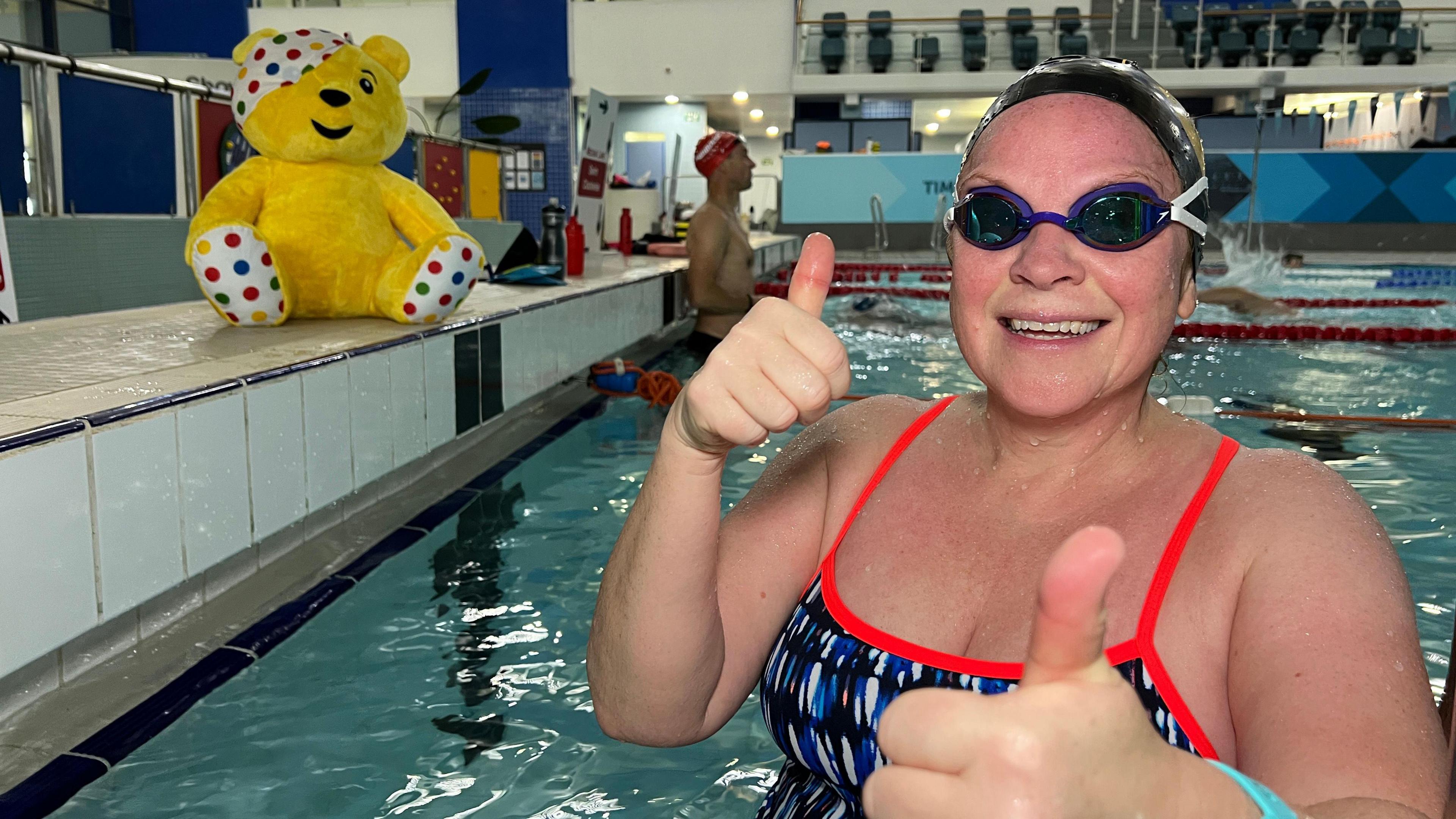 Alison smiles at the camera as she gives a thumbs up with both hands while in the swimming pool. Her goggles are over her eyes and her hair is tucked into her black swim cap. She's wearing a swim costume with a bright red strap over her shoulders. A few people behind her are swimming and there is a Pudsey bear toy by the pool.