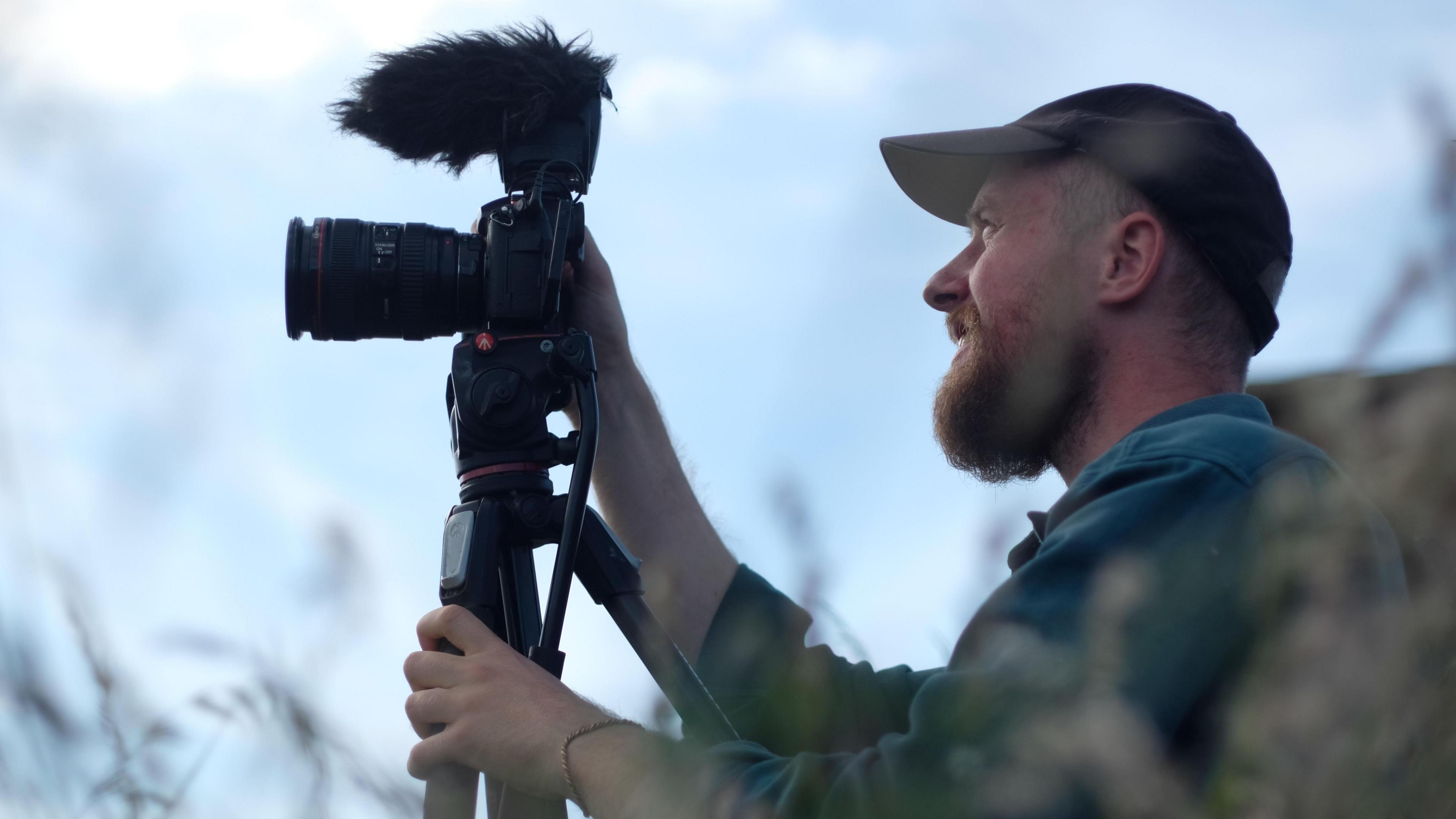 Matthew is wearing a green long sleeved jumper and black baseball cap. He's holding and looking at the screen on his digital camera which is resting on a tripod.