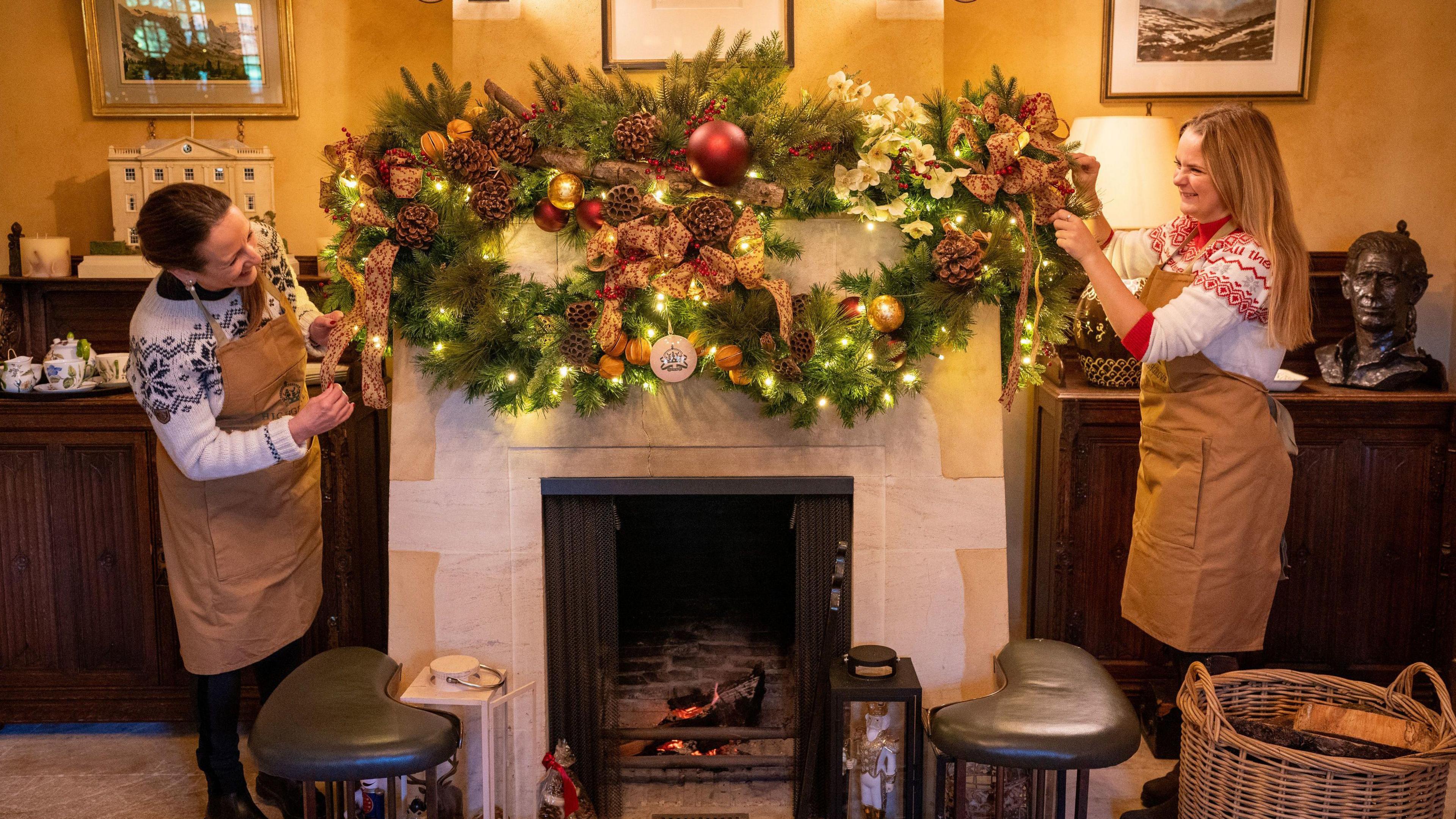 Two Highgrove staff stand either side of a fireplace laying Christmas wreaths on top of it, with fairy lights inside them. There is a small fire in the fireplace