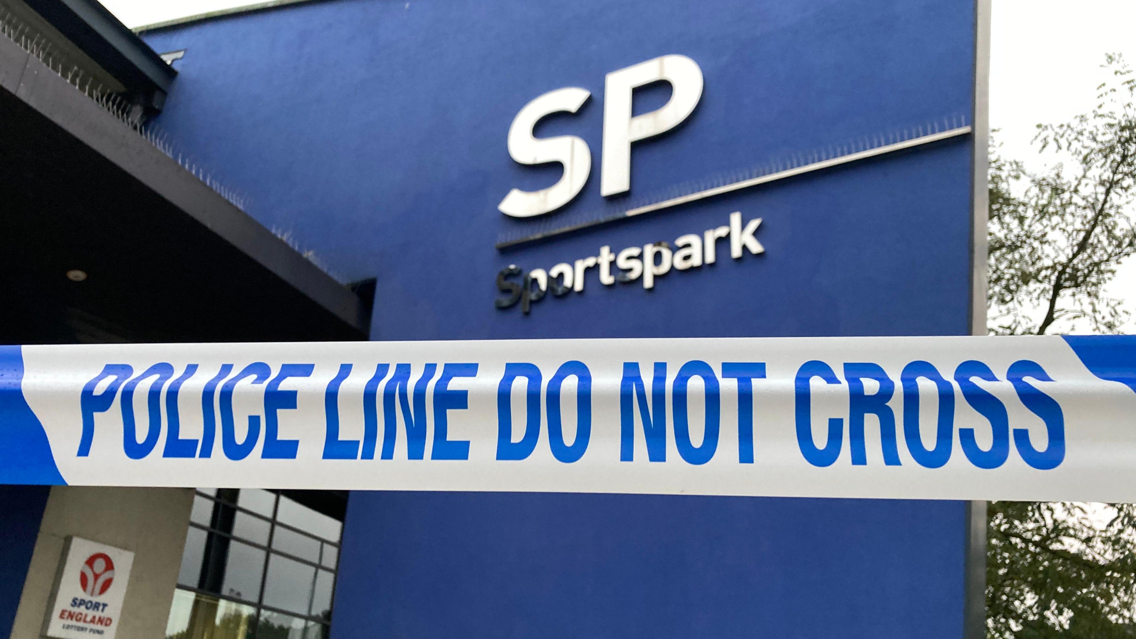 Police tape in front of the Sportspark building. The part of the building in vision is a large blue wall with the silver lettering "SP" and "Sportspark".