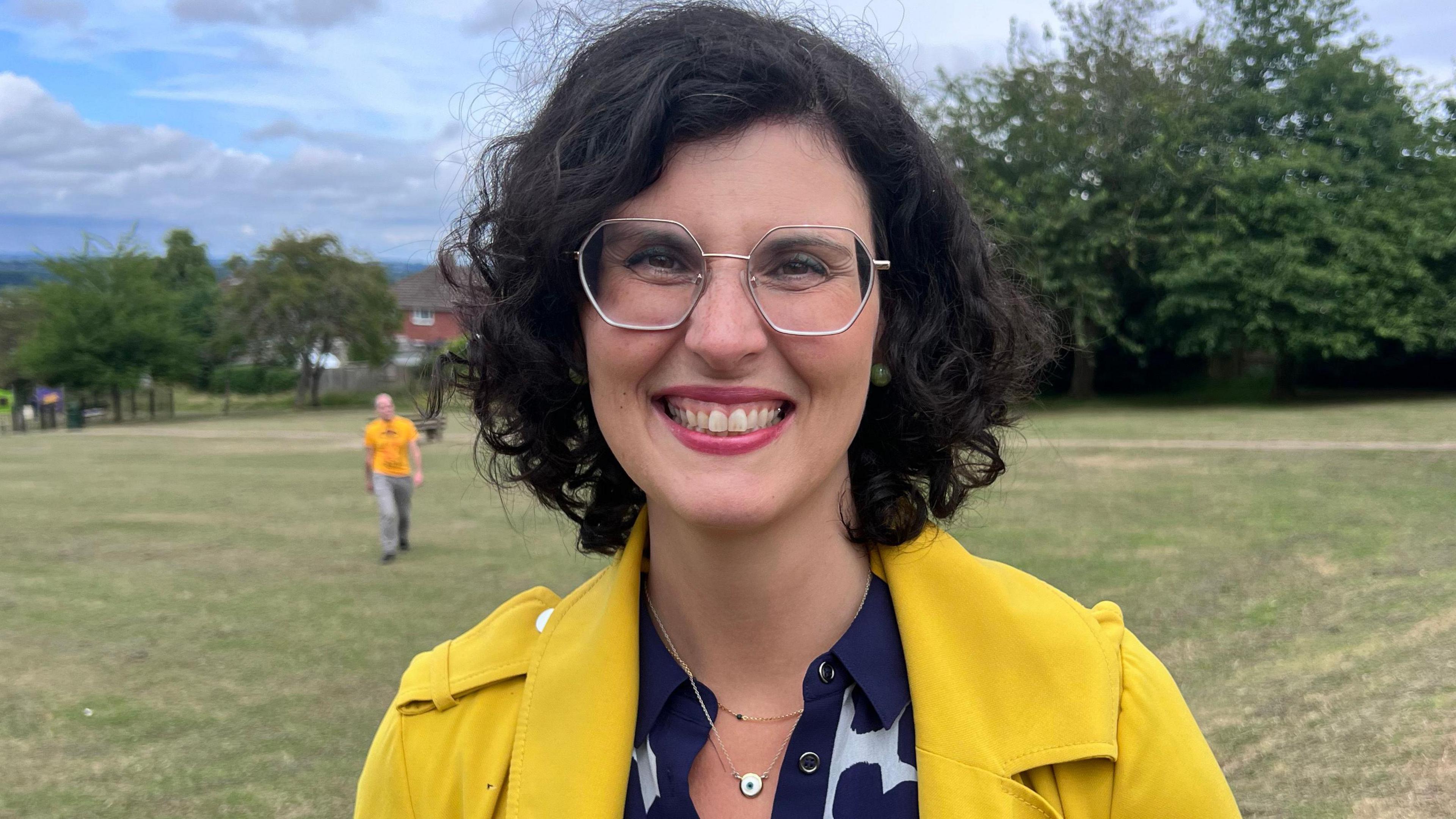 Layla Moran standing in a field wearing a yellow coat and blue and whit shirt. She has medium length brown hair and wears glasses.