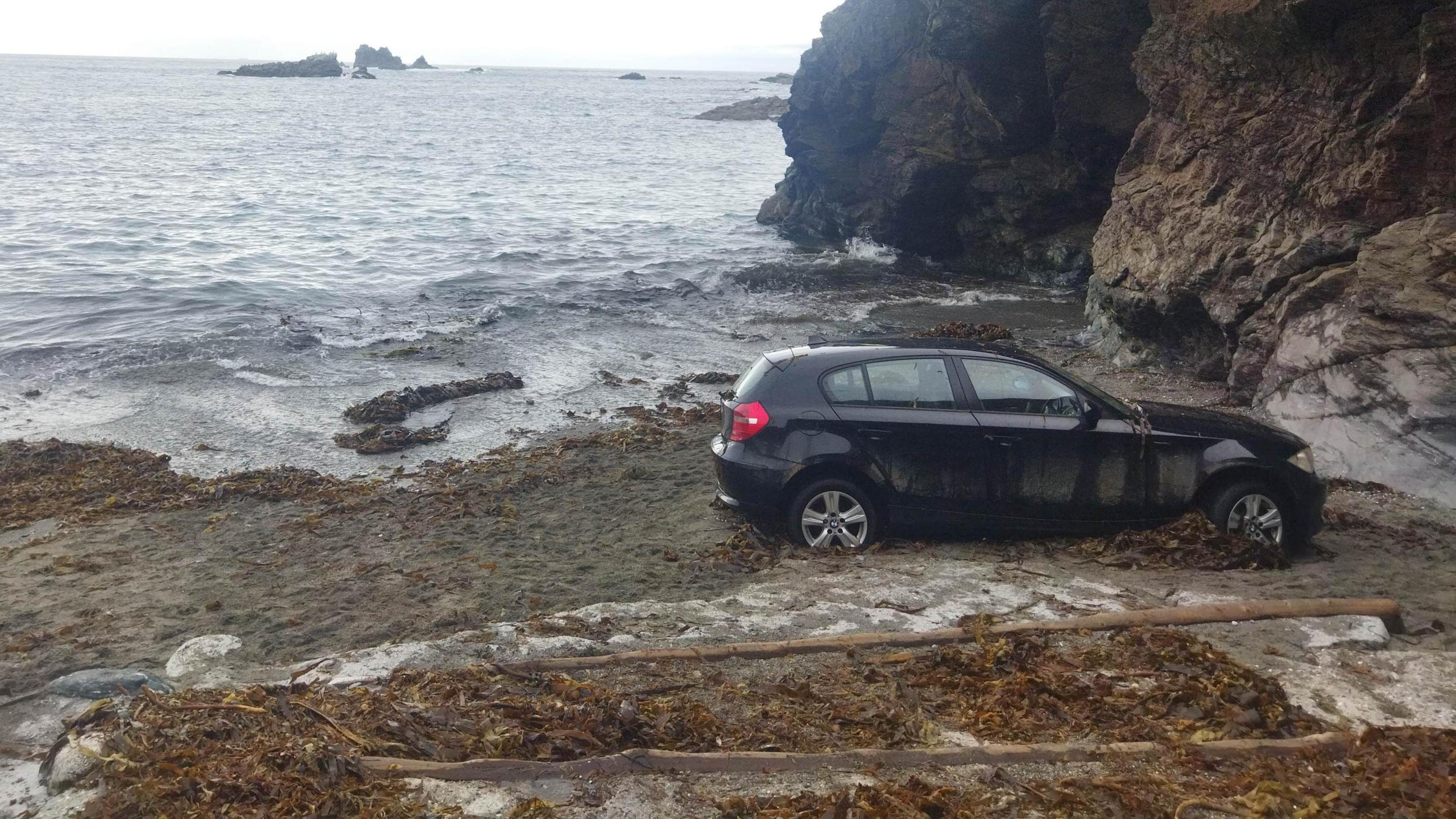 The black BMW is parked facing and about a metre away from the rocky side of the cover. The waves are about five metres from the car and the beach is sandy and has seaweed on it. The last section of slipway is visible in the foreground.