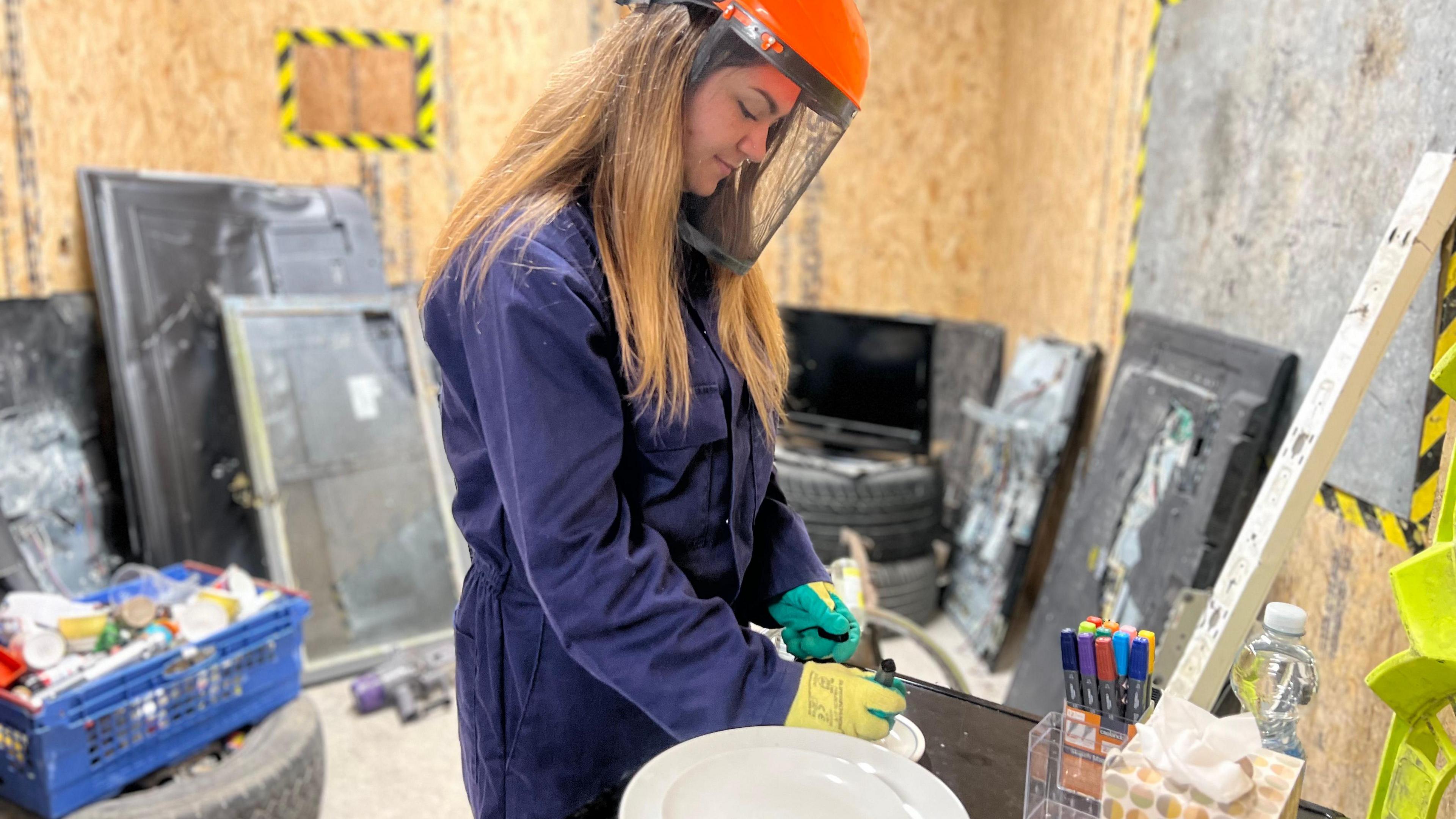 Ashleigh Cowling writing on a plate. She is wearing blue overalls and has safety gear on. In the background is smashed up items and a crate full of bottles and crockery. 