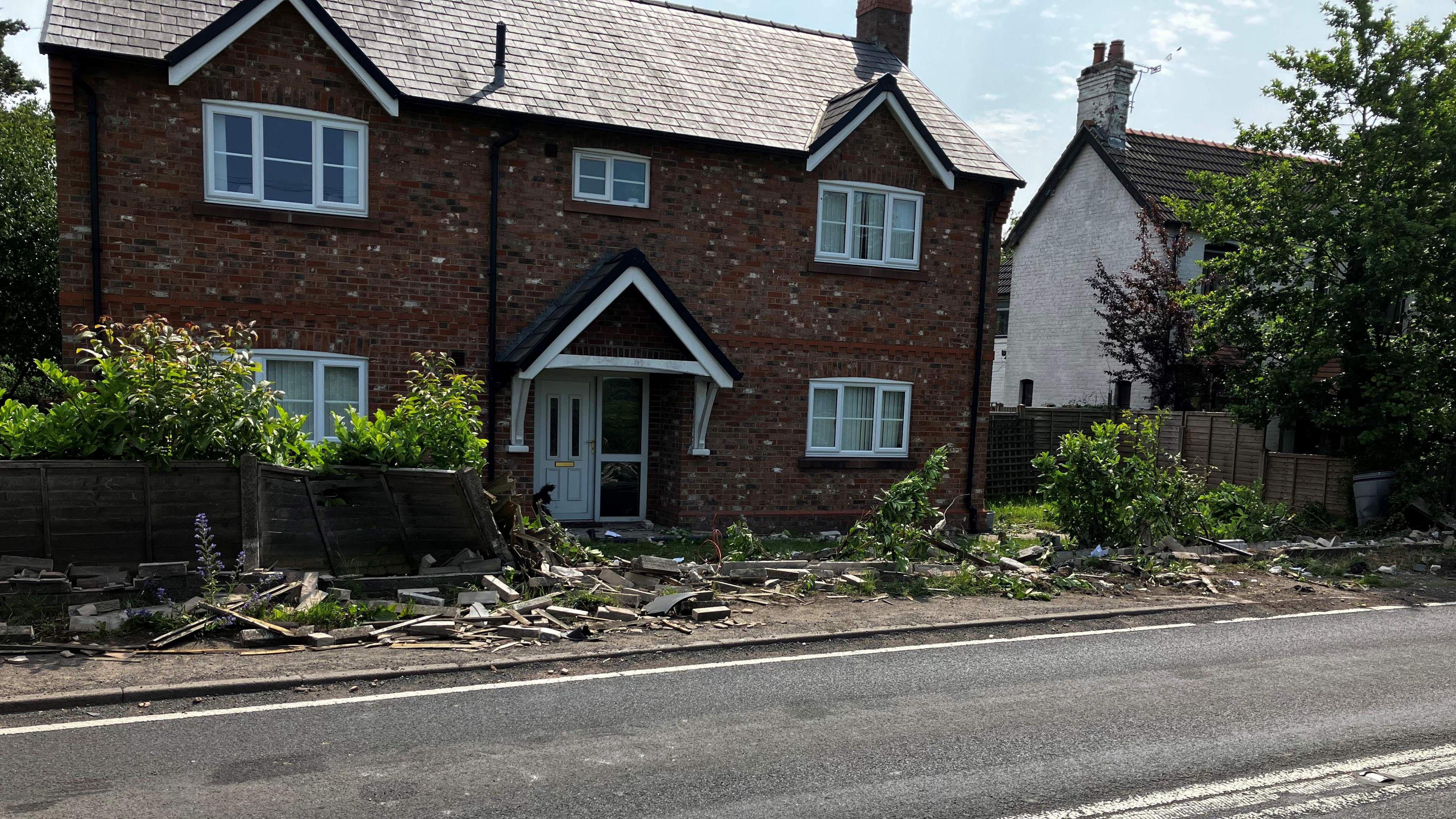 A detached two-storey house set back from a main road. The brick wall and fence in front of it has been destroyed, with rubble lying everywhere.