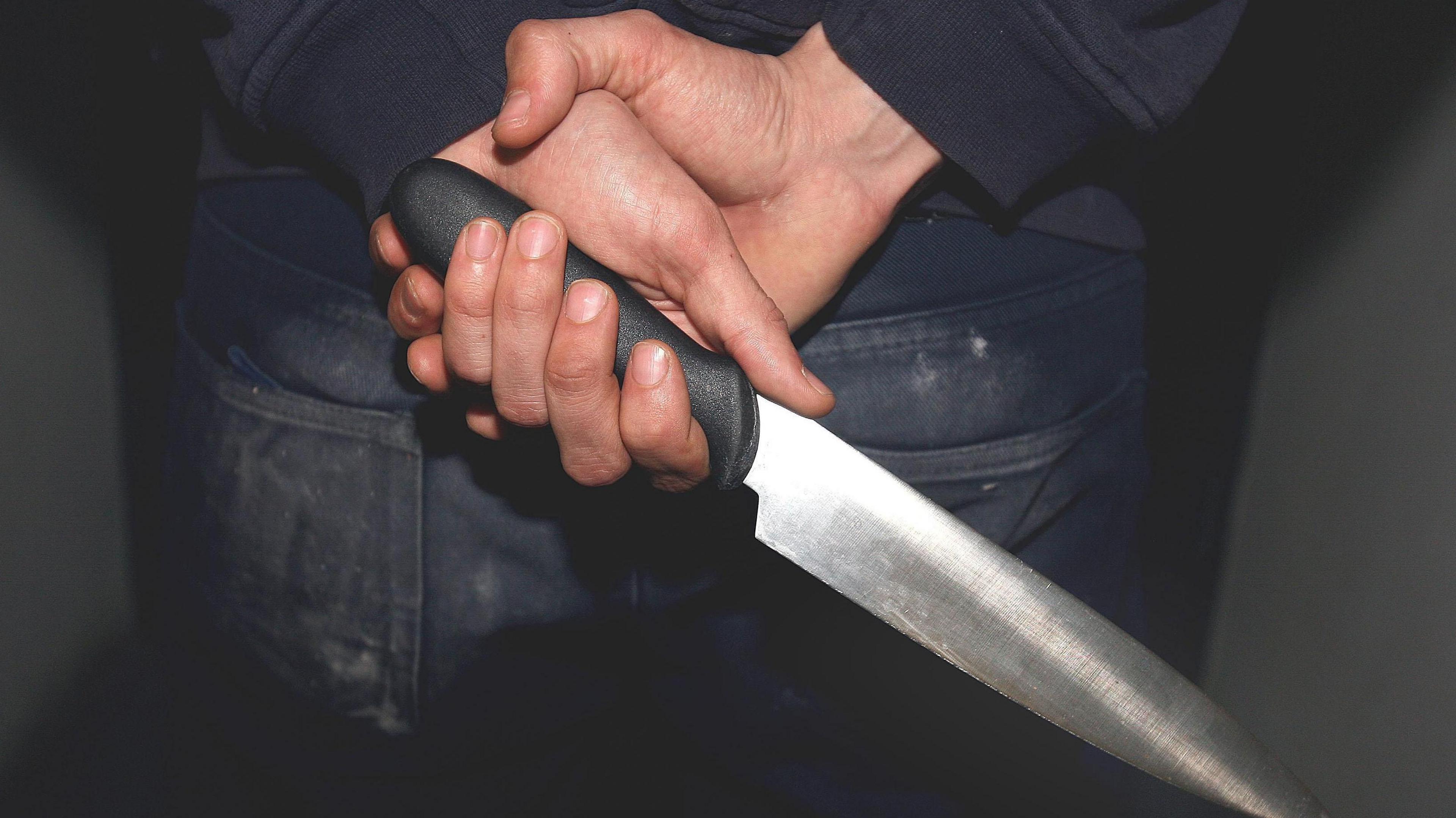 A close-up image of a person holding a large knife behind their back. They are wearing a navy jumper with navy jeans. The knife has a black hilt