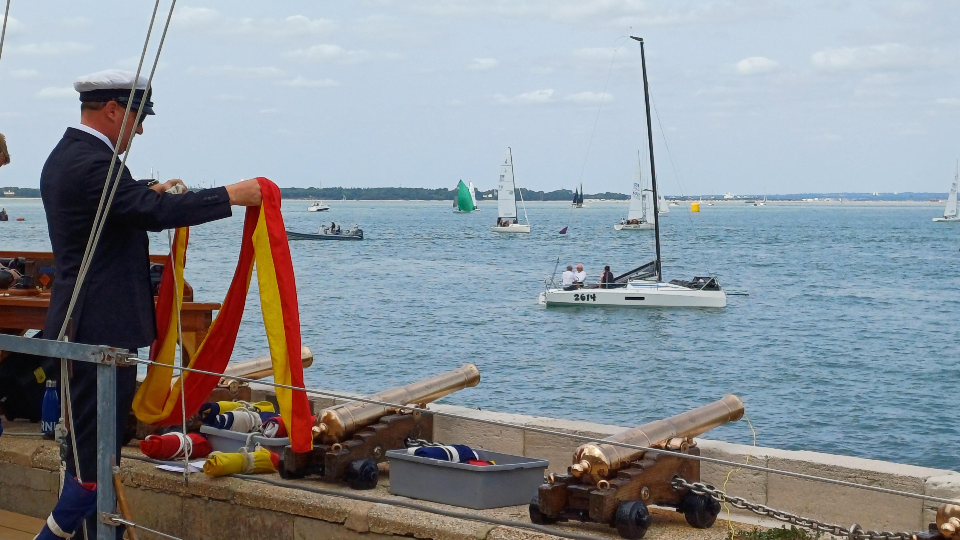 A man in a nautical blazer and cap unfurls a red and yellow flag. Three small canons sit on a way in front of him. A sea view dominated the remainder of the image with some small yachts in full sail and one nearer the camera with it's sails stowed