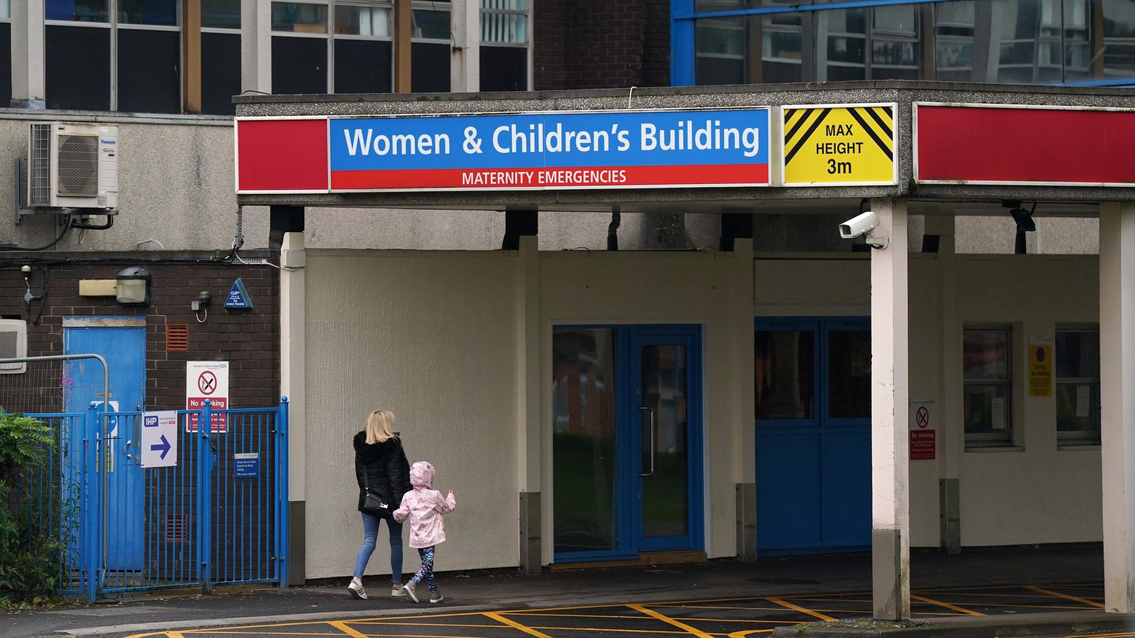 The entrance to the Women and Children's Building at the Countess of Chester Hospital. A blonde woman in a black coat walks under a sign for the building hand-in-hand with a young girl in a pink coat. 