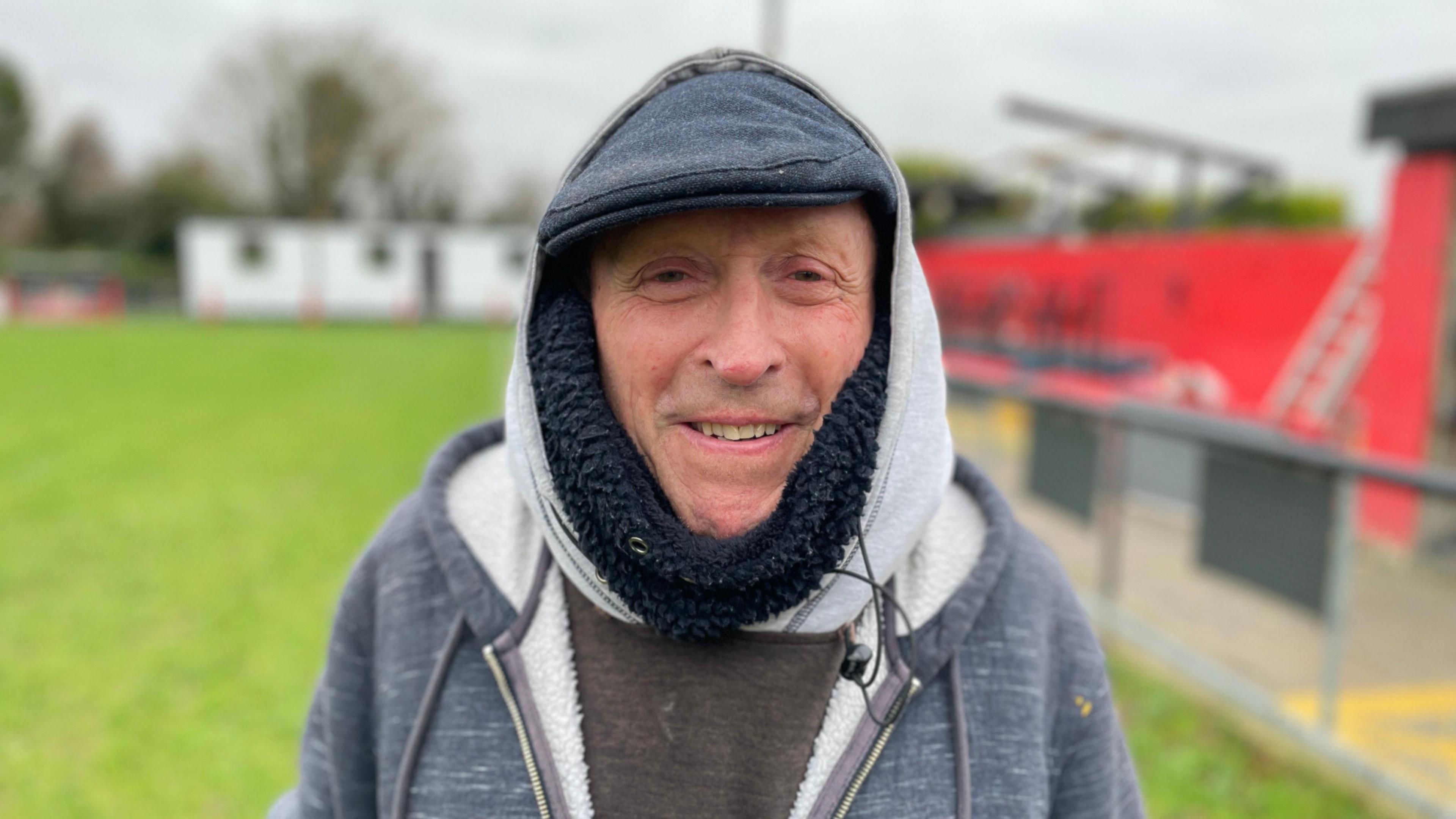 Steve Day standing on a football pitch wearing a hoodie and a cap. He is wearing a jumper underneath his hoodie. He is wearing a cap and has the hood of his hoodie over his head.