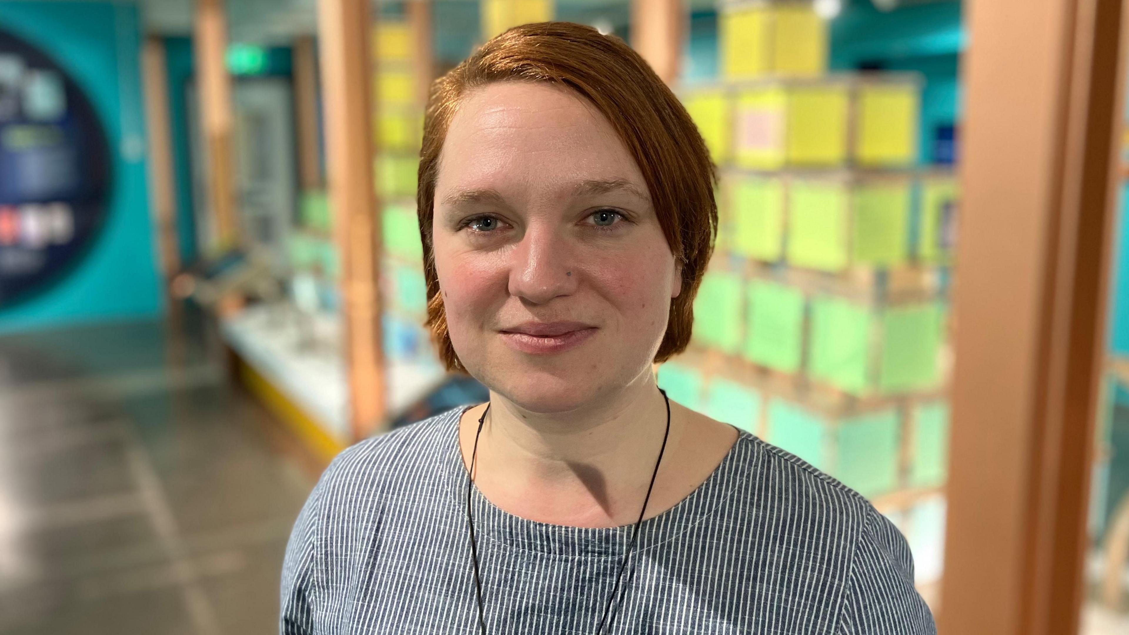 Erica from exhibitions team at Bletchley Park looking directly into the camera with the colourful exhibition behind her. She has ginger hair and is wearing a stripey black and white top.