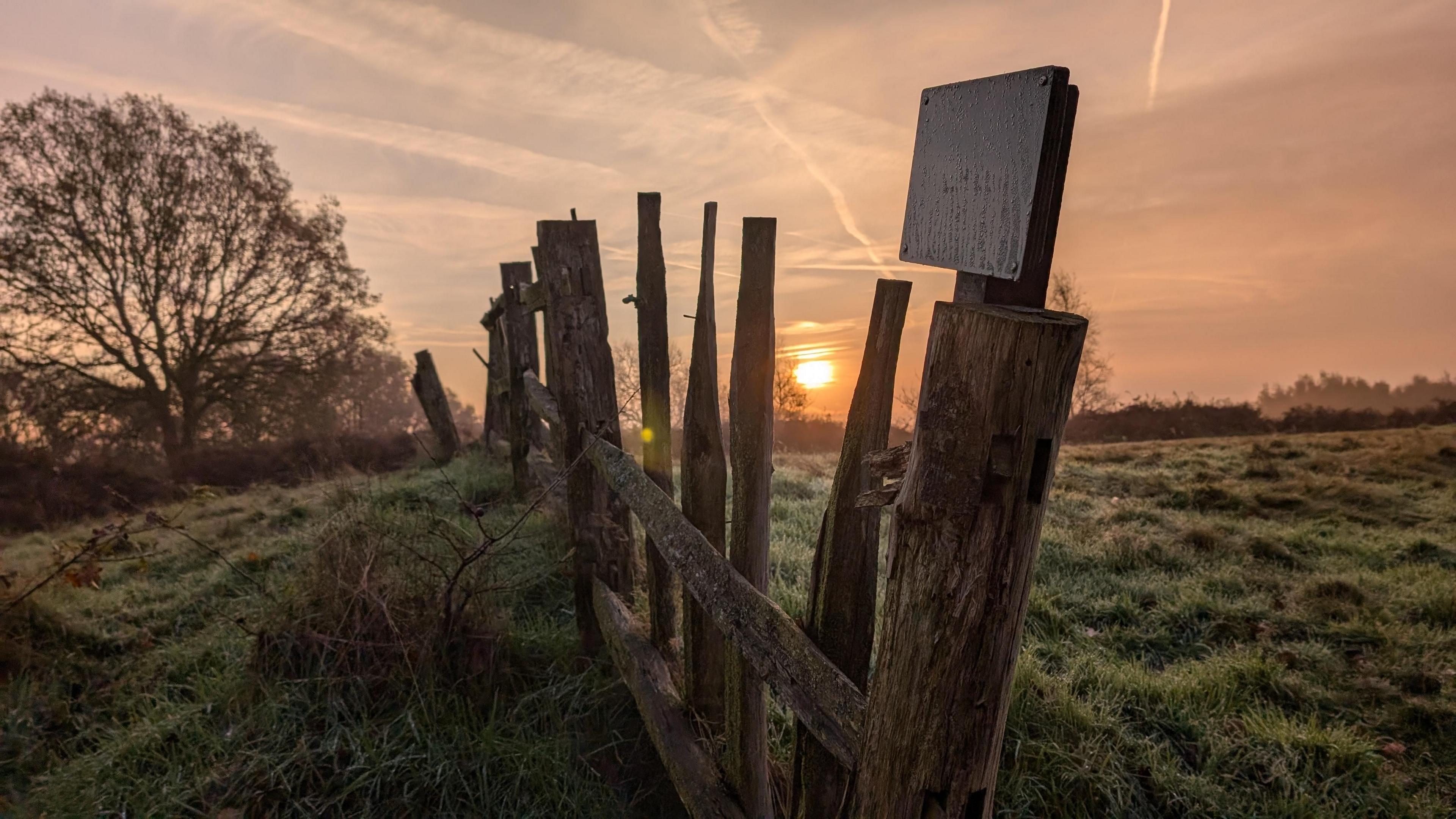 A rough-cut wooden fence with a weather-worn sign on it is in the foreground of a shot with a dewy or frosty field and the sun rising over the horizon.