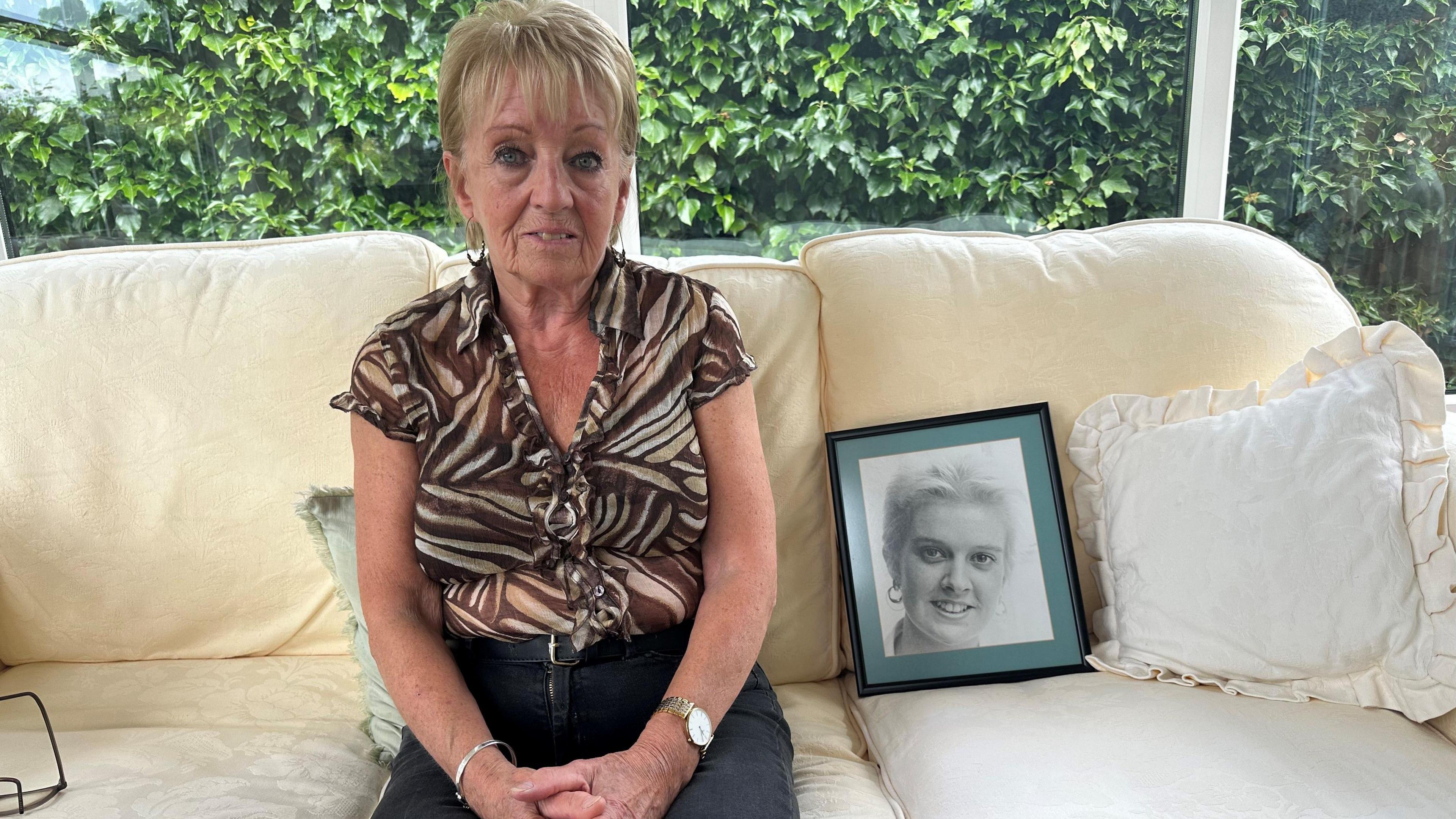 Doreen Soulsby sitting on a sofa next to a picture of her daughter. She's wearing a smart brown shirt and black trousers 