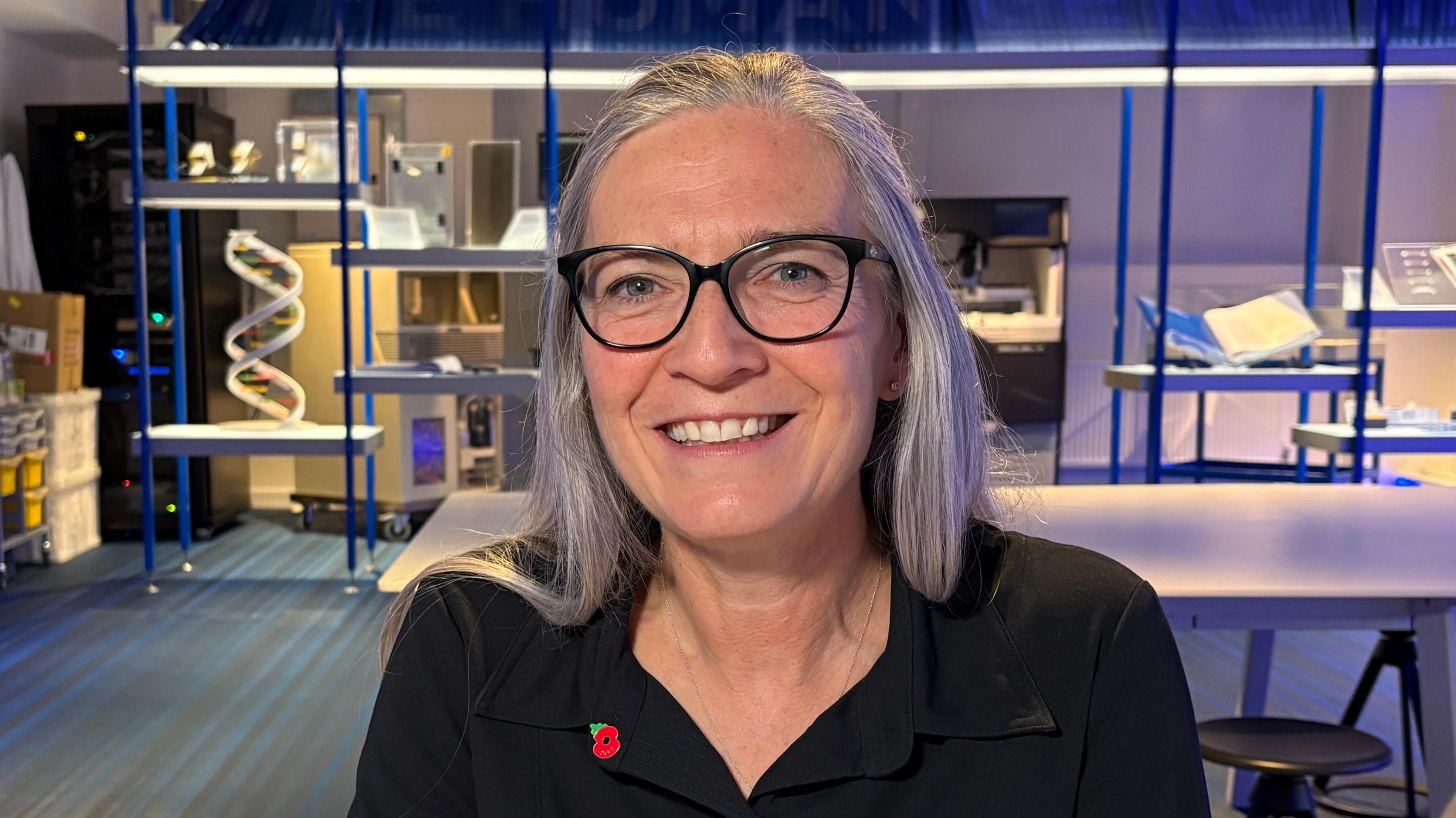 Dr Julia Wilson is looking towards the camera smiling. She has long grey hair and is wearing blacked framed glasses, a black shirt and a poppy badge on her collar. 