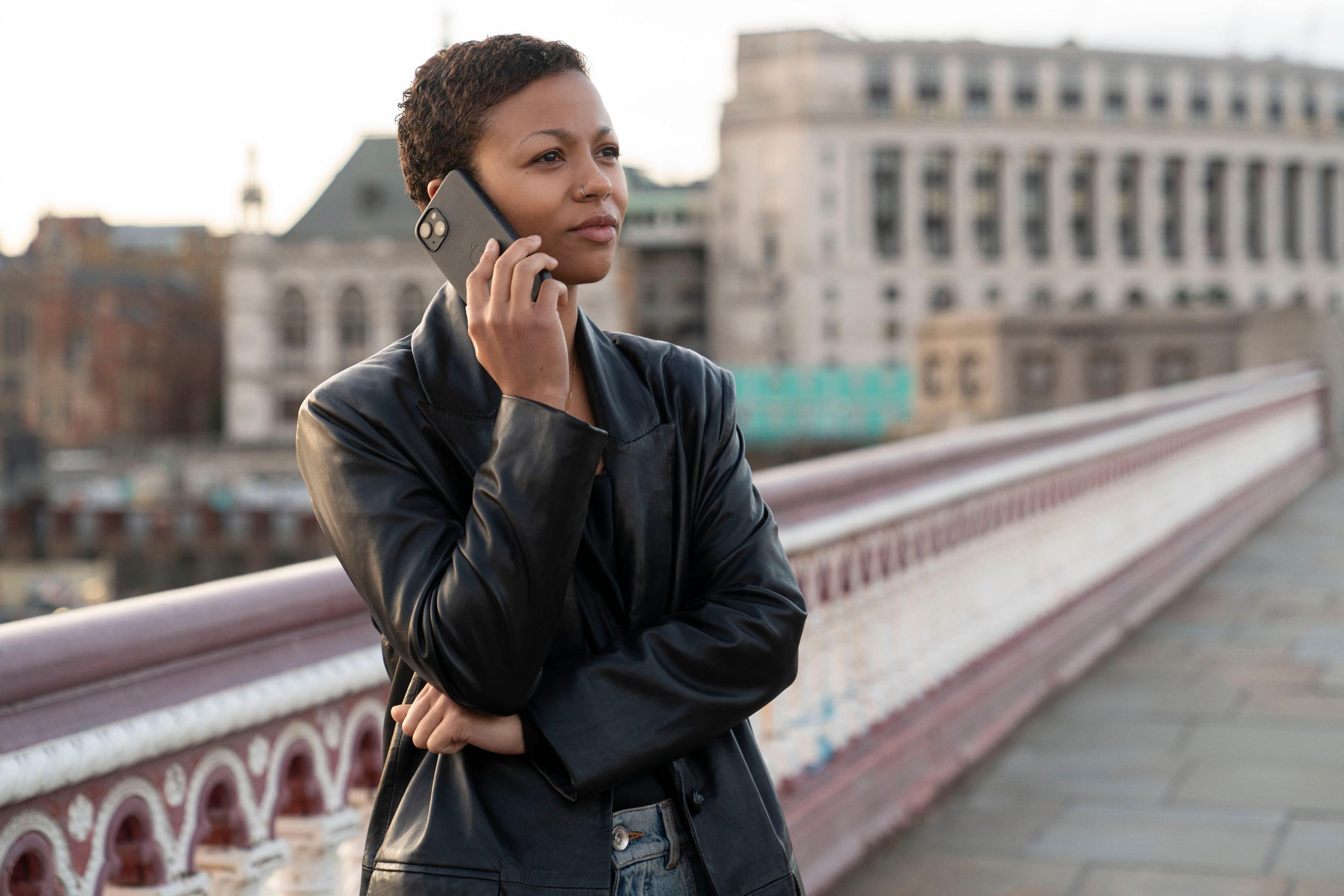 A woman is dressed with a black leather coat on a bridge, with a phone to her ear