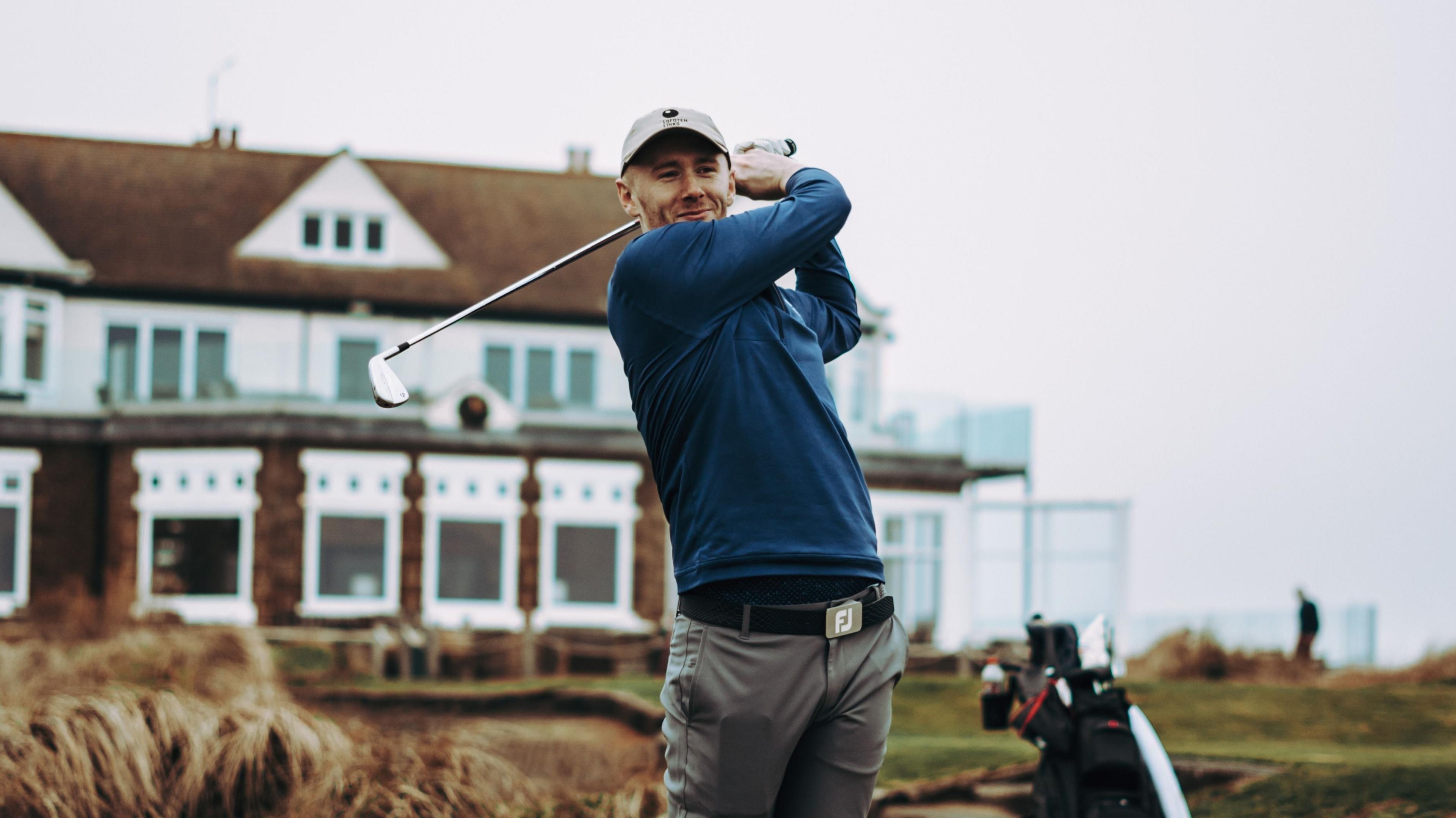 Golfer Josh Simpson stood with his club after teeing off with a clubhouse behind him in the distance.
