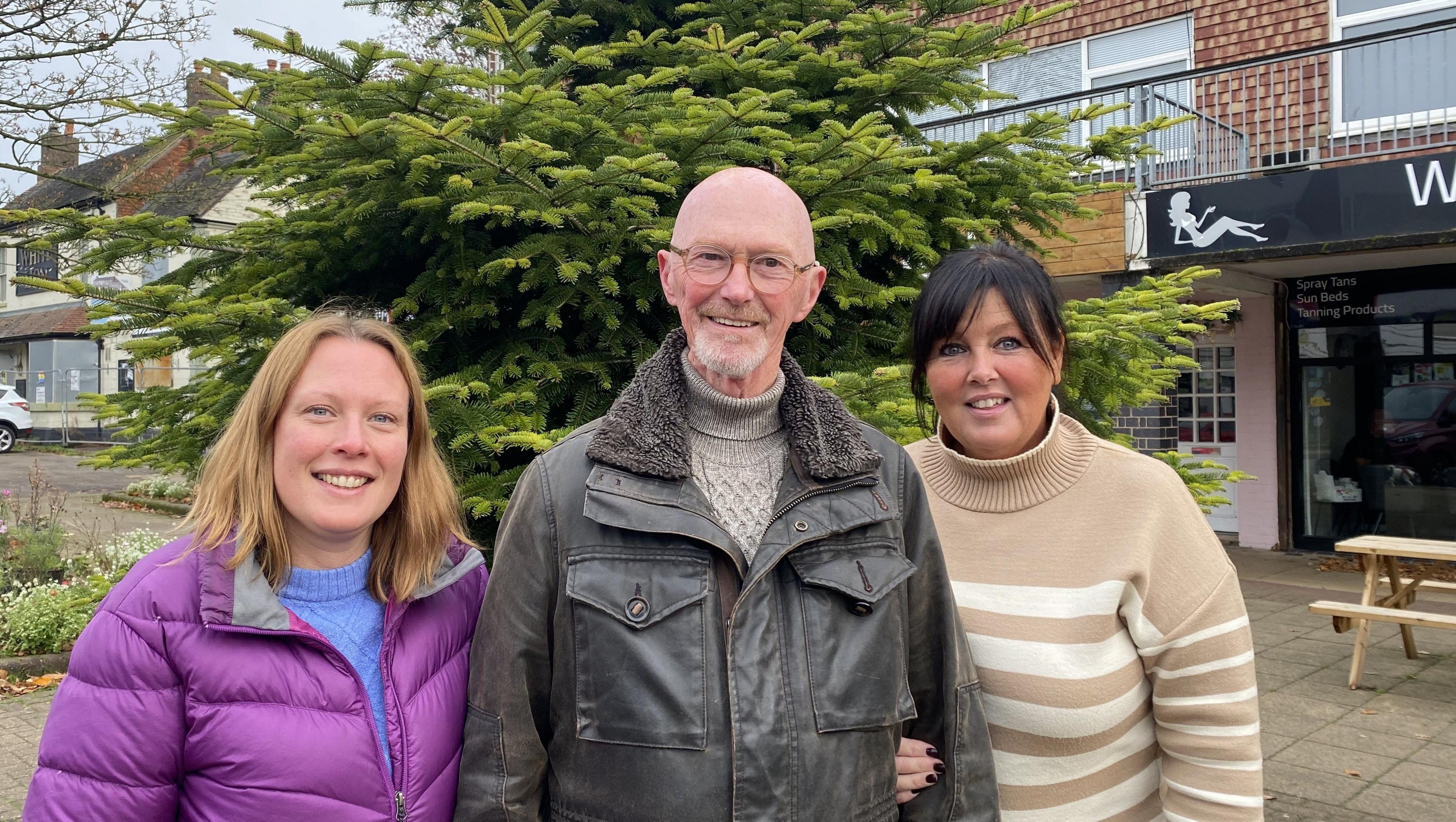 Ruth Ramshaw in a purple coat, Richard Smith in a leather jacket and a hairdresser in a cream stripy jumper stood in front of a Christmas tree 