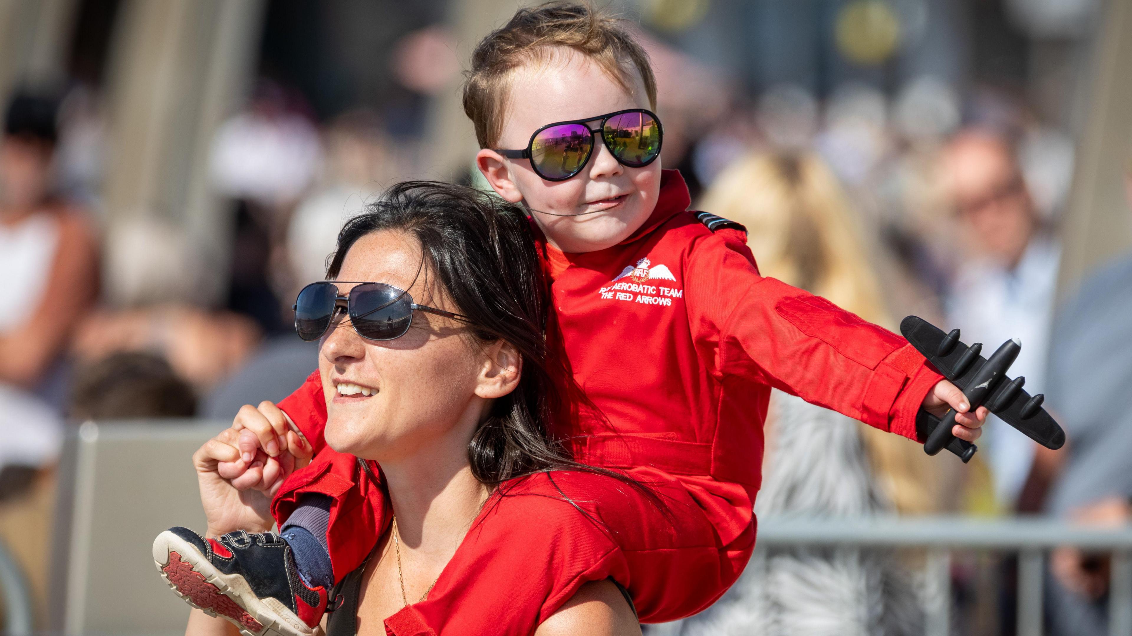 A young boy in a Red Arrows red jumpsuit and mirrored sunglasses waves a toy plane as he sits on a woman's shoulders