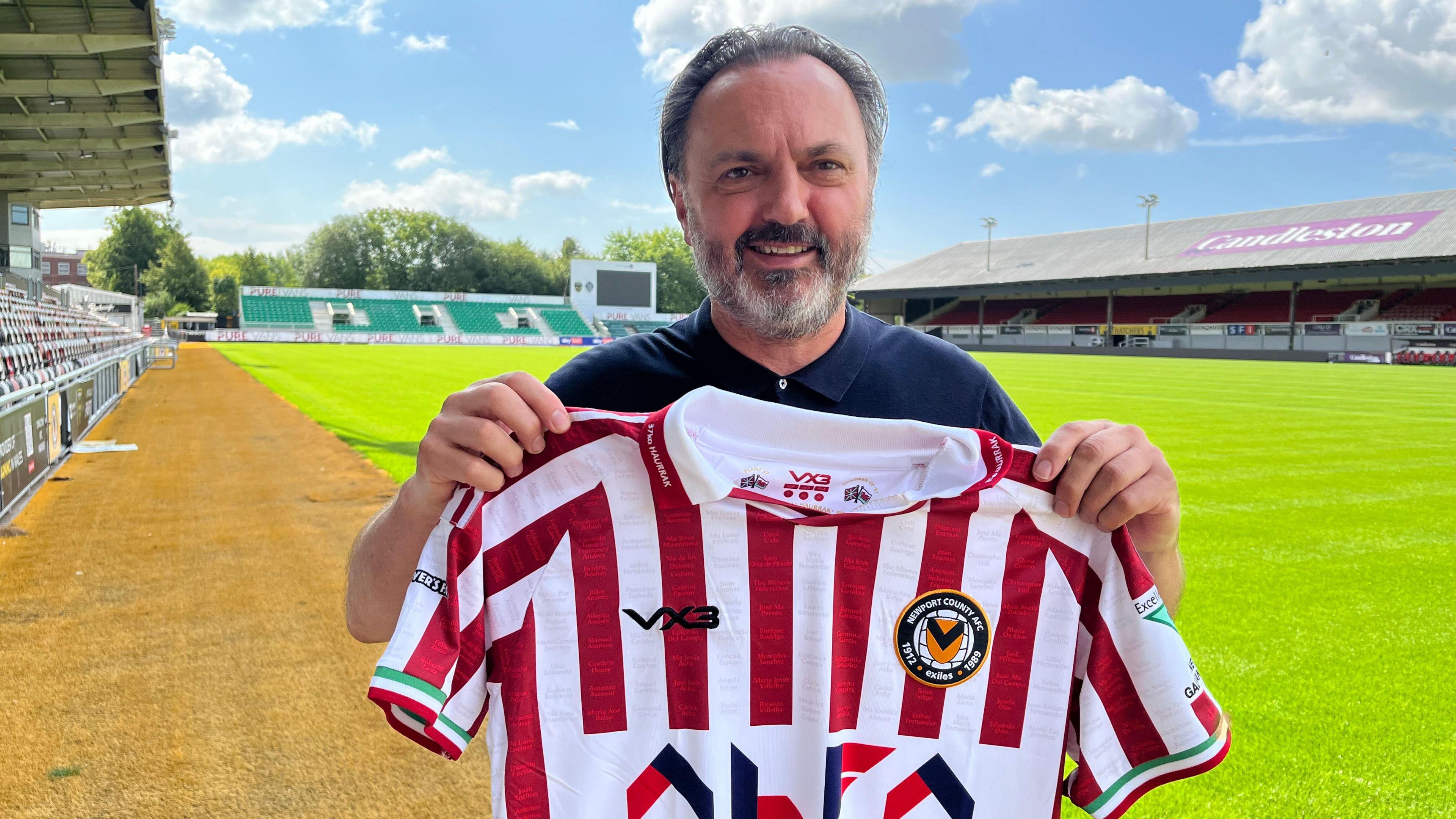 Neal Heard with the Newport County 2025/26 away kit. The shirt which is a red and white striped football shirt. He is stood on the football pitch, the green grass is behind and to the left of him and the football stands surround him. It is a sunny day.