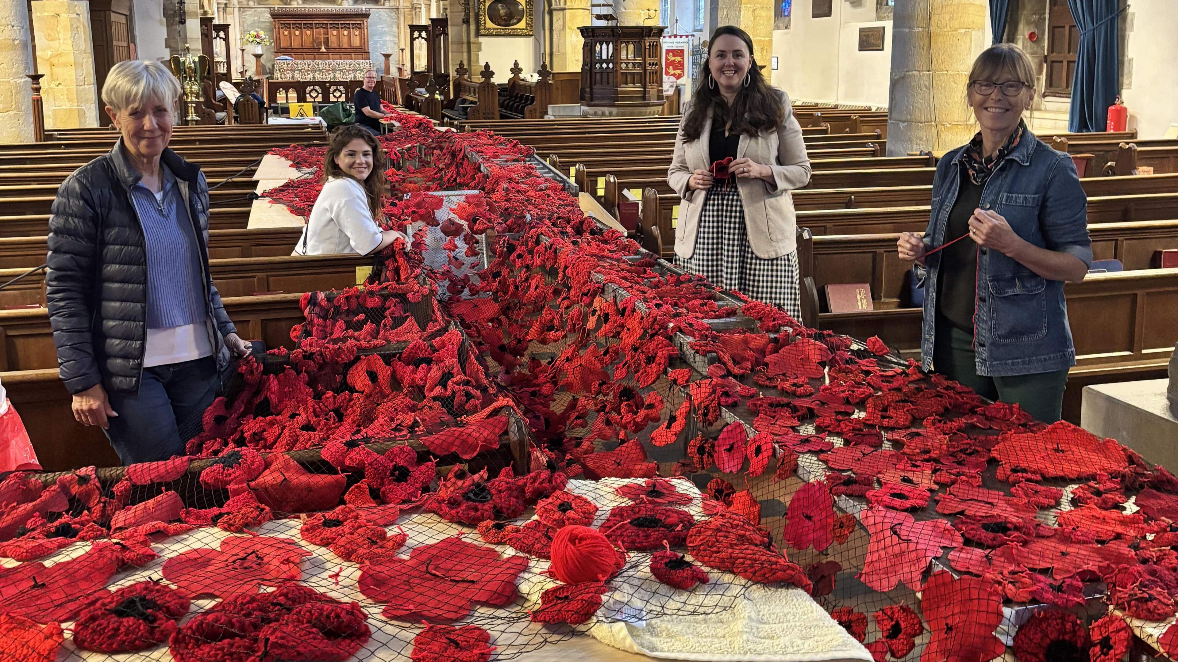 Four women smiling at the camera while standing in a church. In the aisle there are hundreds of handmade poppies.