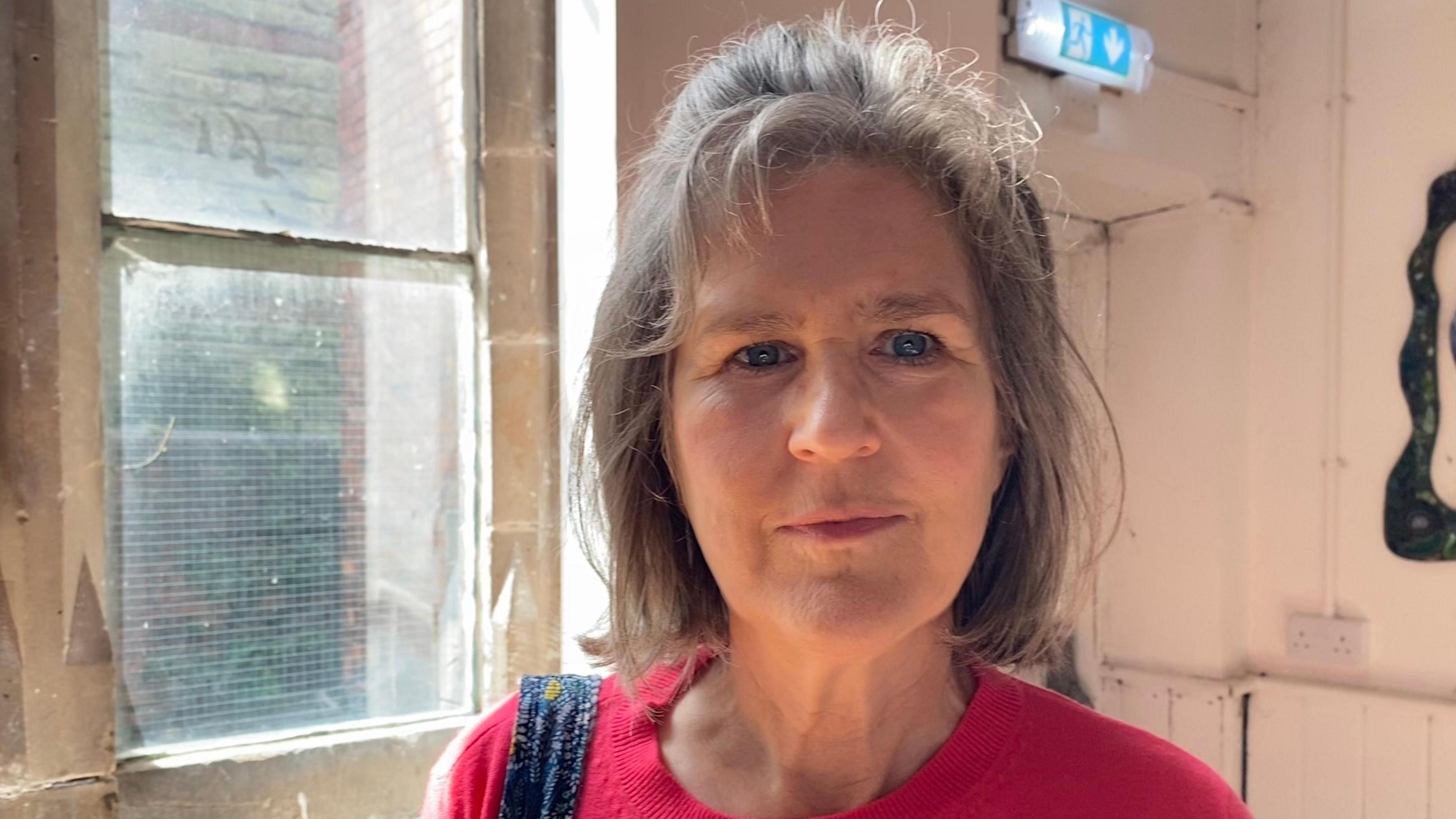 A woman with shoulder length light brown hair is standing in a room with a window behind her. She is wearing a red jumper.