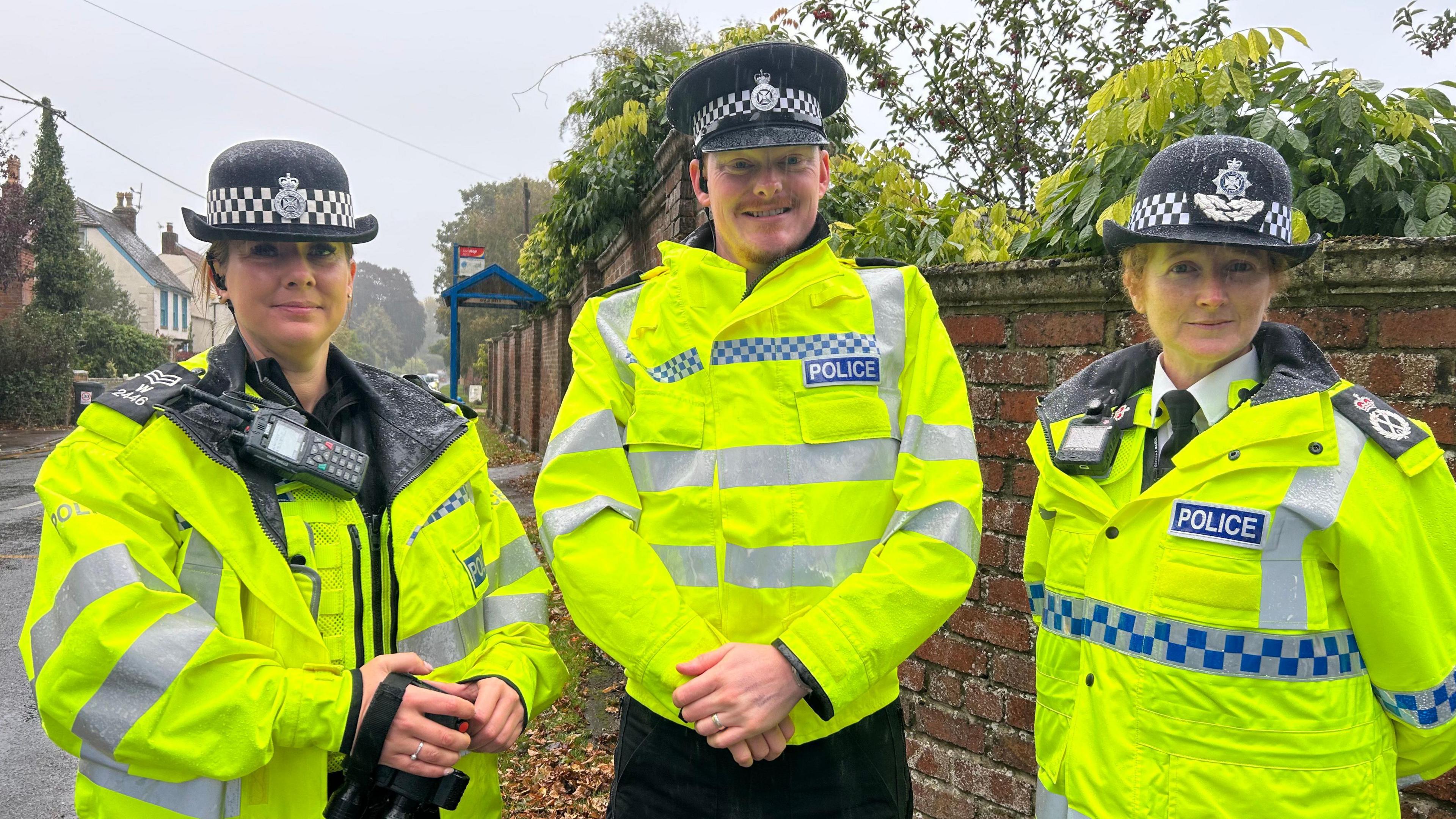 Three police officers wearing uniform and high vis jackets stand together on the pavement.