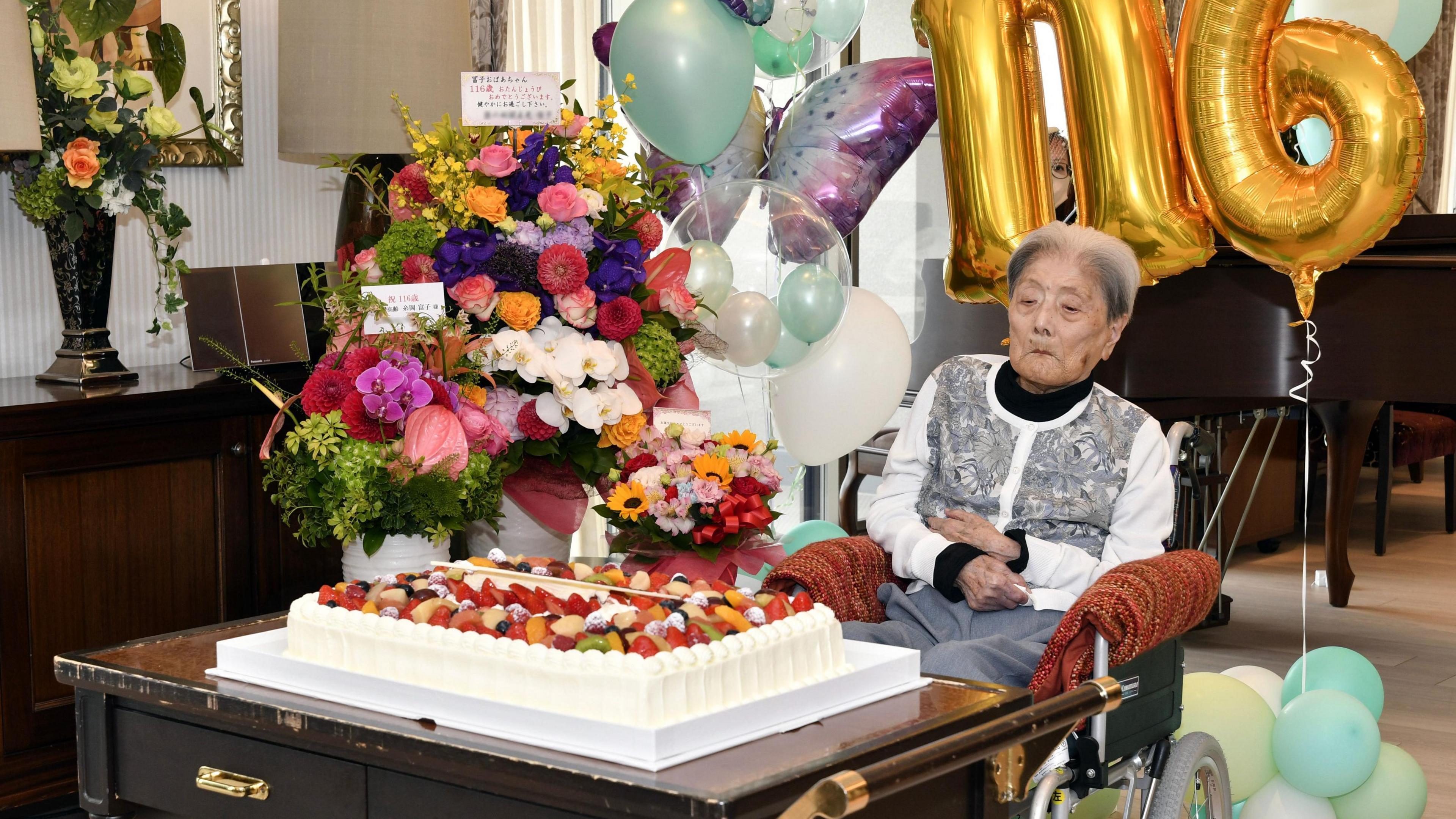 Tomiko Itooka celebrates her 116th birthday. She sits in front of a large white cake topped with fruit and a bouquet of flowers. Gold helium ballons in the shapes of 116 loiter behind her. 