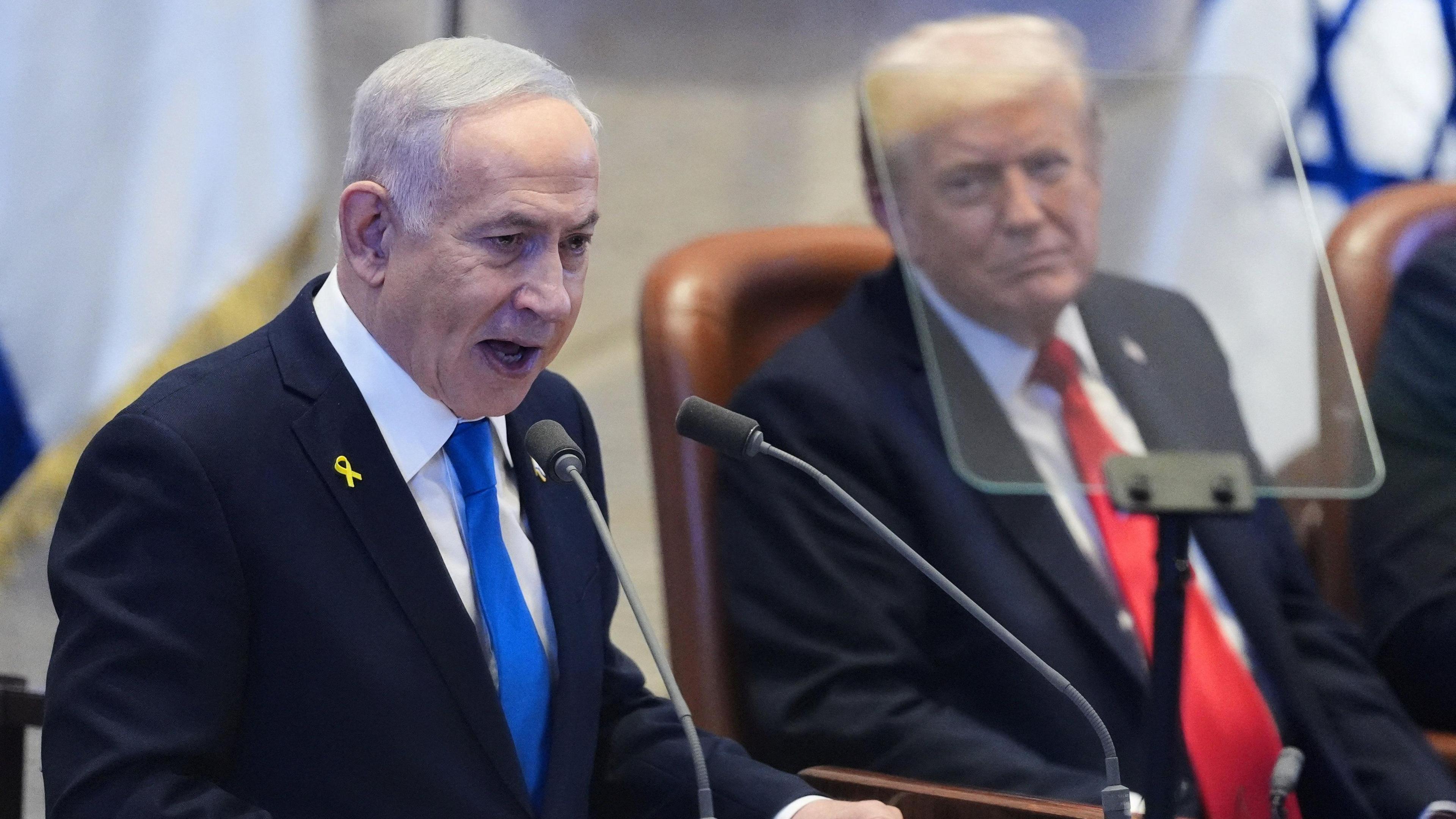 Benjamin Netanyahu wears a suit and blue tie, standing in front of a microphone. President Trump is in the background wearing a red tie and a suit.