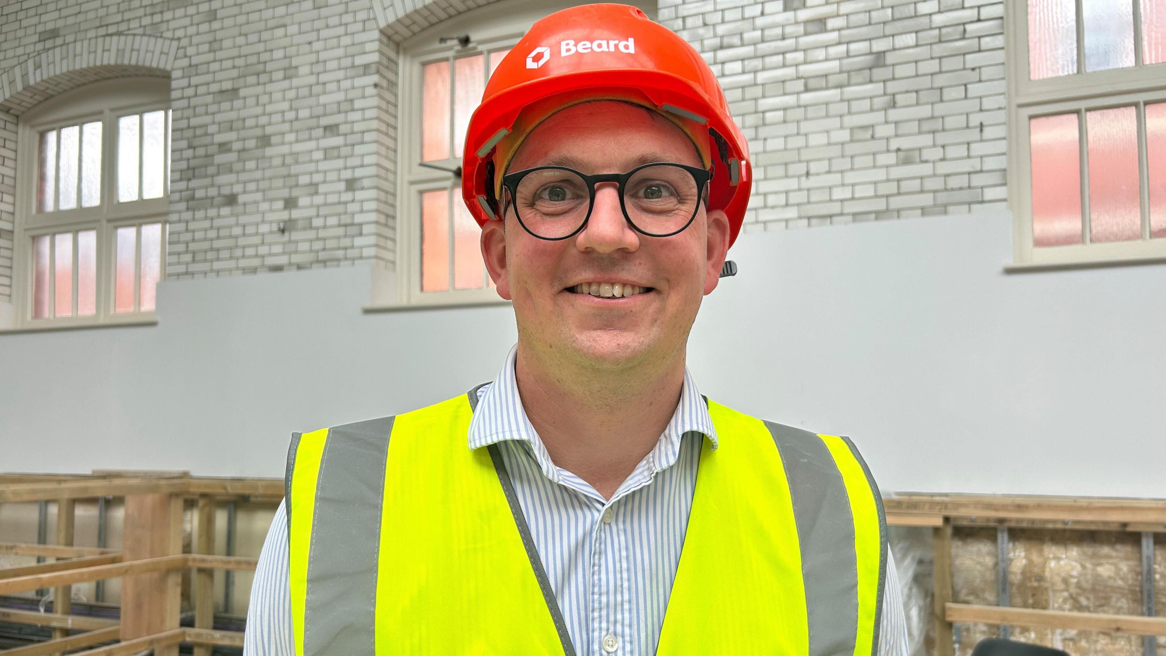 The project lead is smiling wearing a blue and white striped shirt, black rounded glasses, a yellow high vis jacket and bright orange hard hat with the company name, Beard written on the front. Behind him are the white glazed bricks in the gym and insulation which is being fitted into the walls.