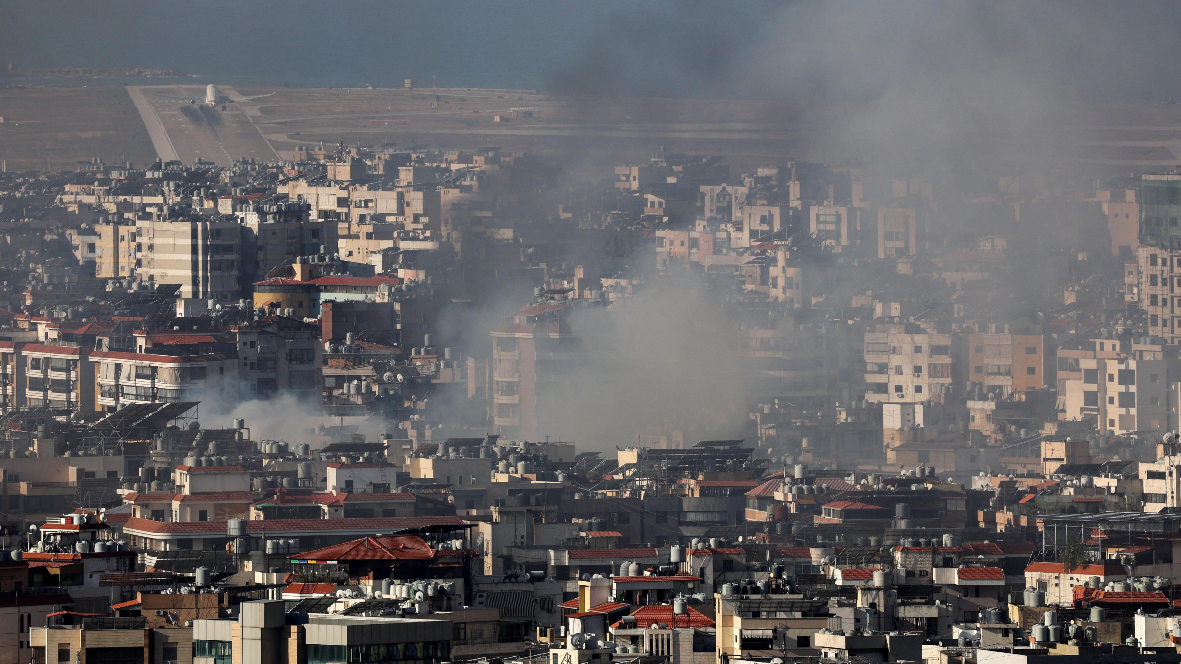 A plane takes off from a runway in the background as smoke rises from a city in the foreground.