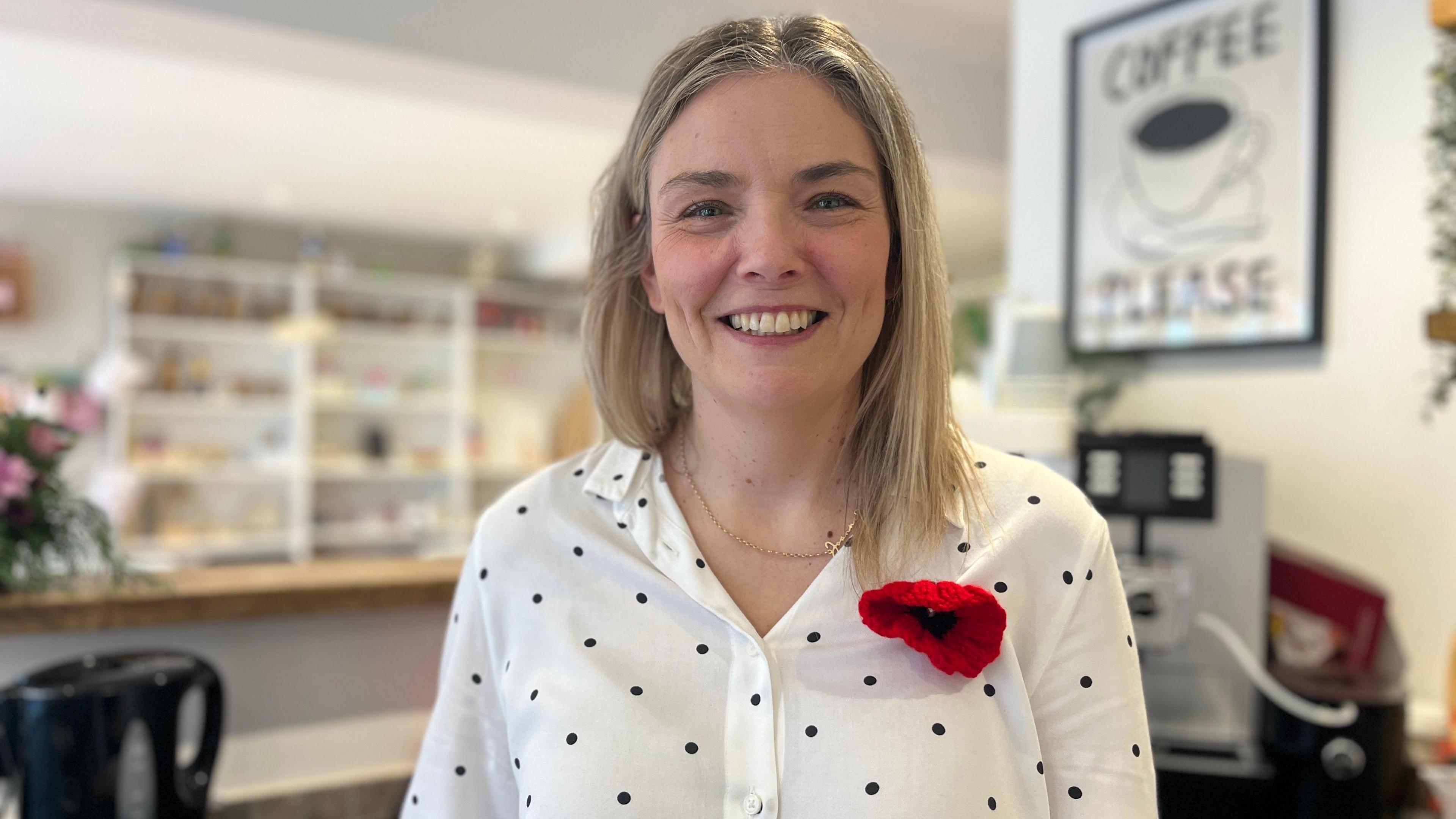 A woman with blonde hair is smiling at the camera. She is wearing a white shirt with black polka dots on it, and has a knitted red and black poppy pinned to her top.