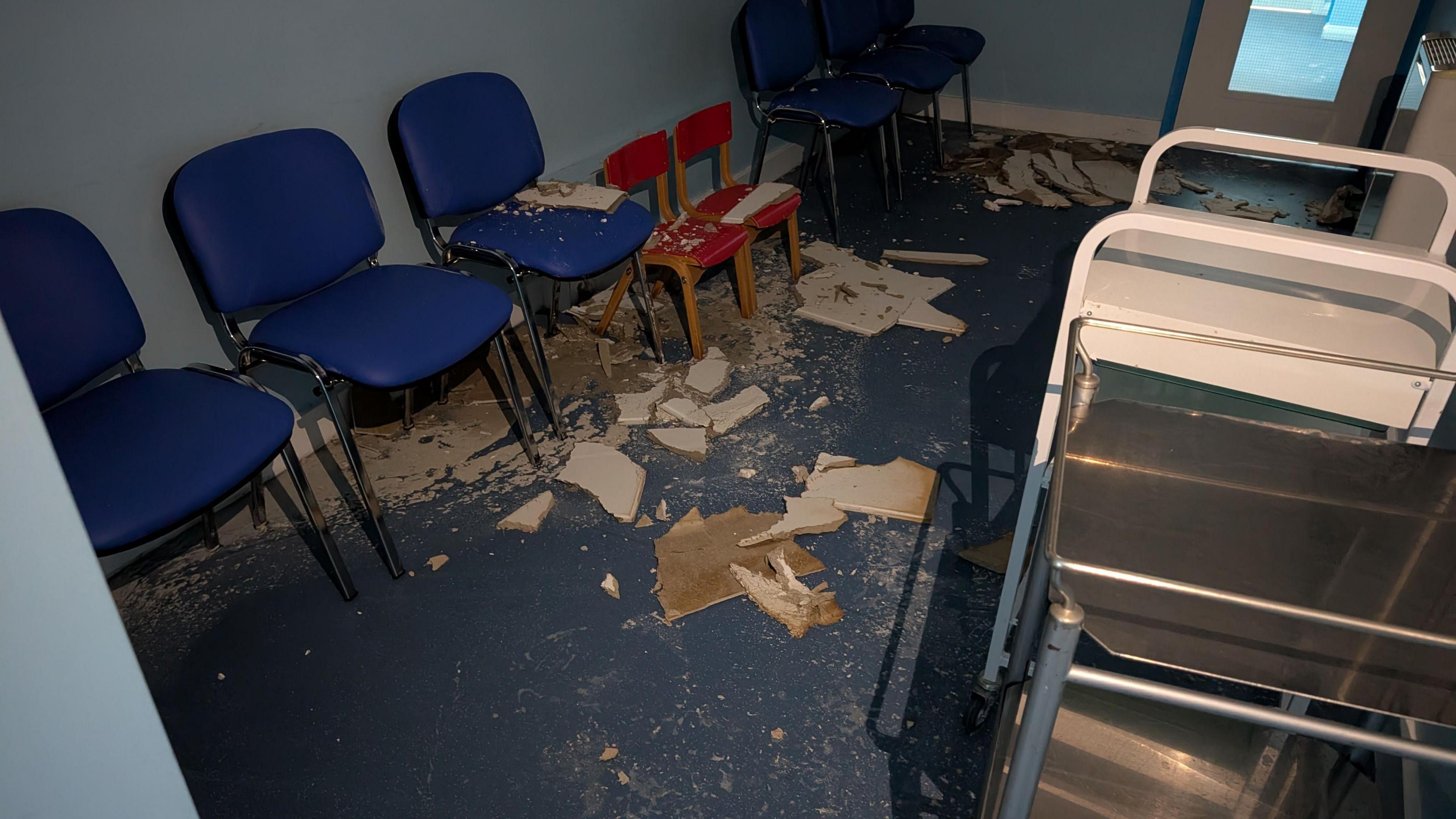 Three large blue chairs and two small red ones are covered in ceiling panels and rubble. 