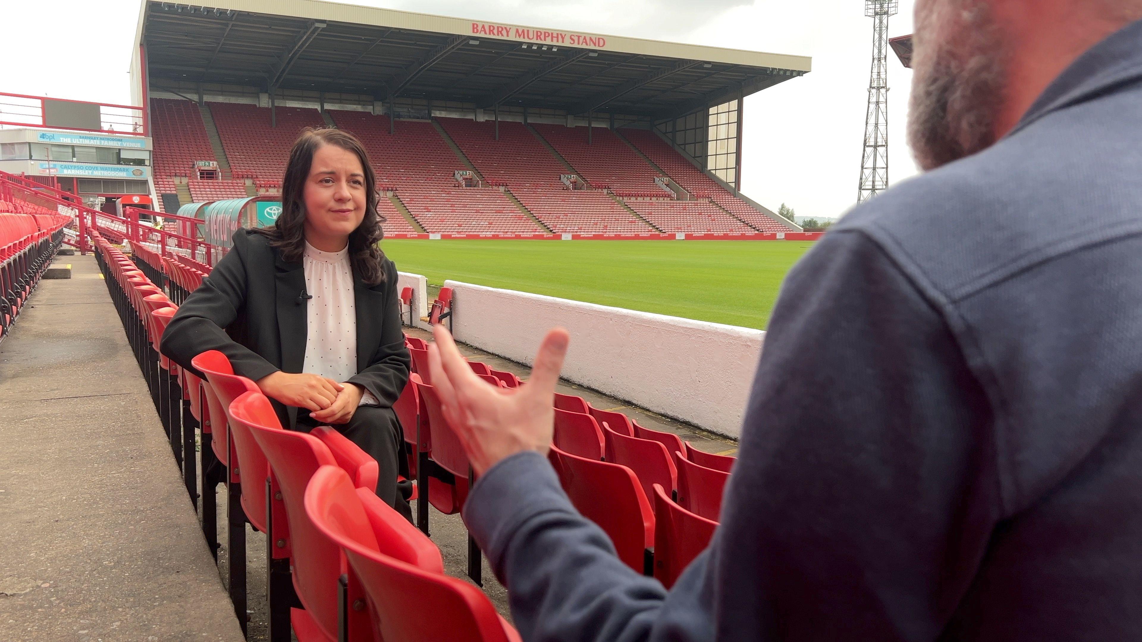 Sports Minster Steph Peacock sits with BBC Yorkshire's James Vincent in Barnsley's Oakwell stadium. She is wearing a white shirt and dark jacket - James Vincent is in the foreground with a blue shirt on. they are surrounded by red seats.
