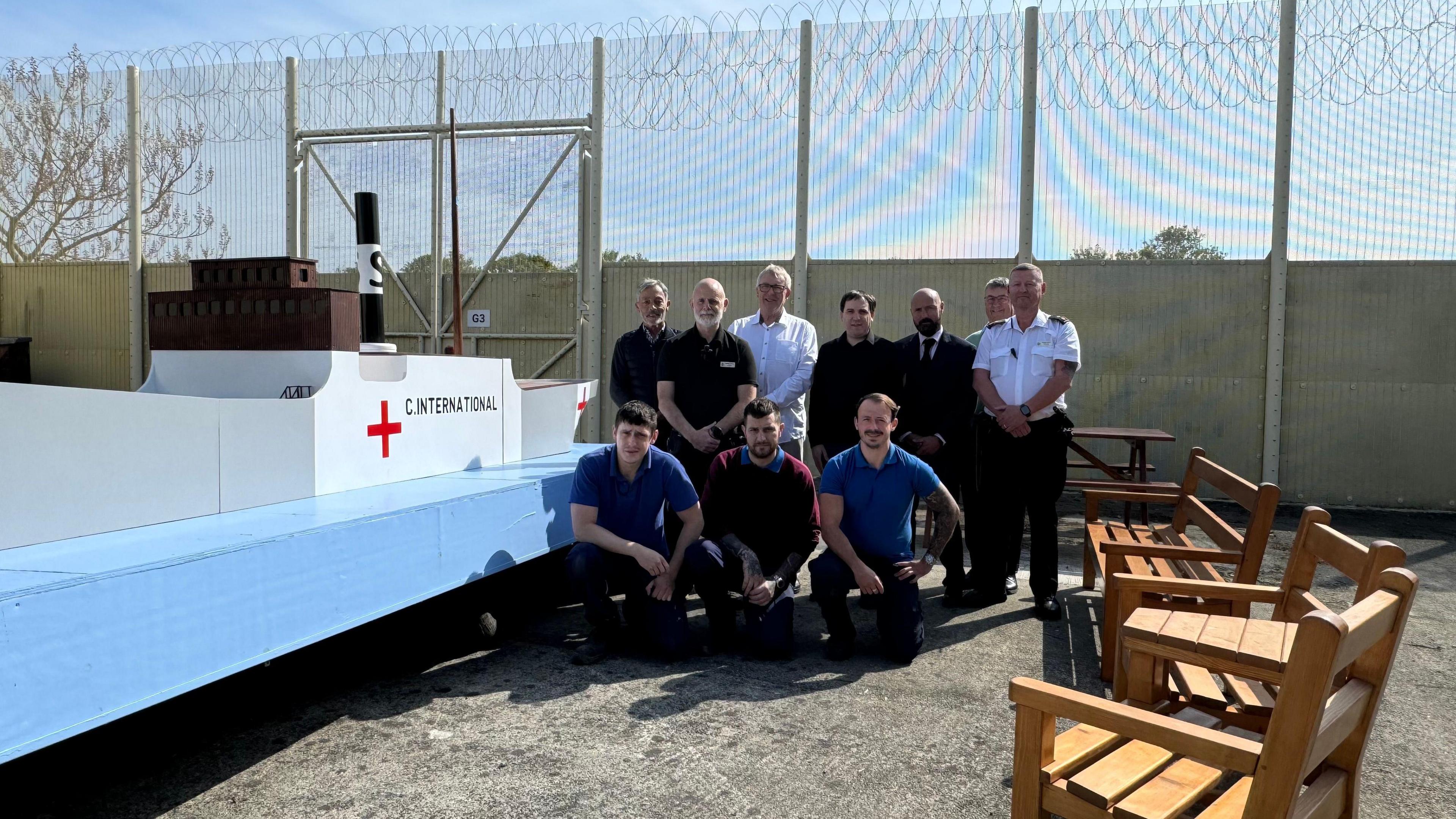 A model of a boat, the SS Vega. It is on a light blue board. The ship is white, with the words C.International on it and a red cross. To the right of the boat are prisoners, kneeling and prison guards stood up and a man in a suit. To the right of them are three benches, faces towards the boat. Behind them is a prison gate. 
