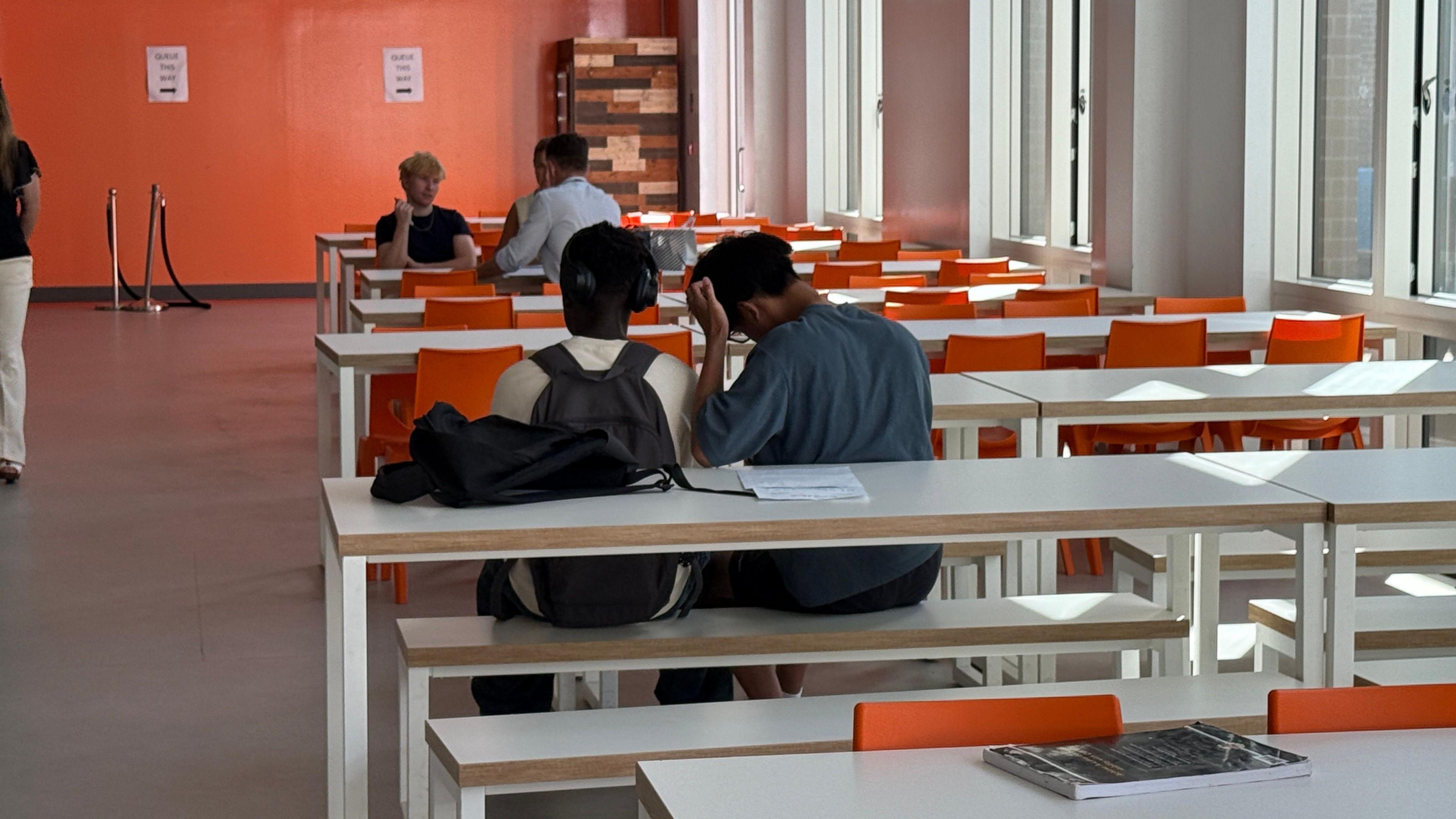 Two men are sat down with their backs to the camera. They are sitting on a white bench at a white table in the canteen. The man on the right is leaning his elbow on the table and has his head propped up on his hand. There is another group of three men a few tables away from the pair, who look like they're having a conversation.