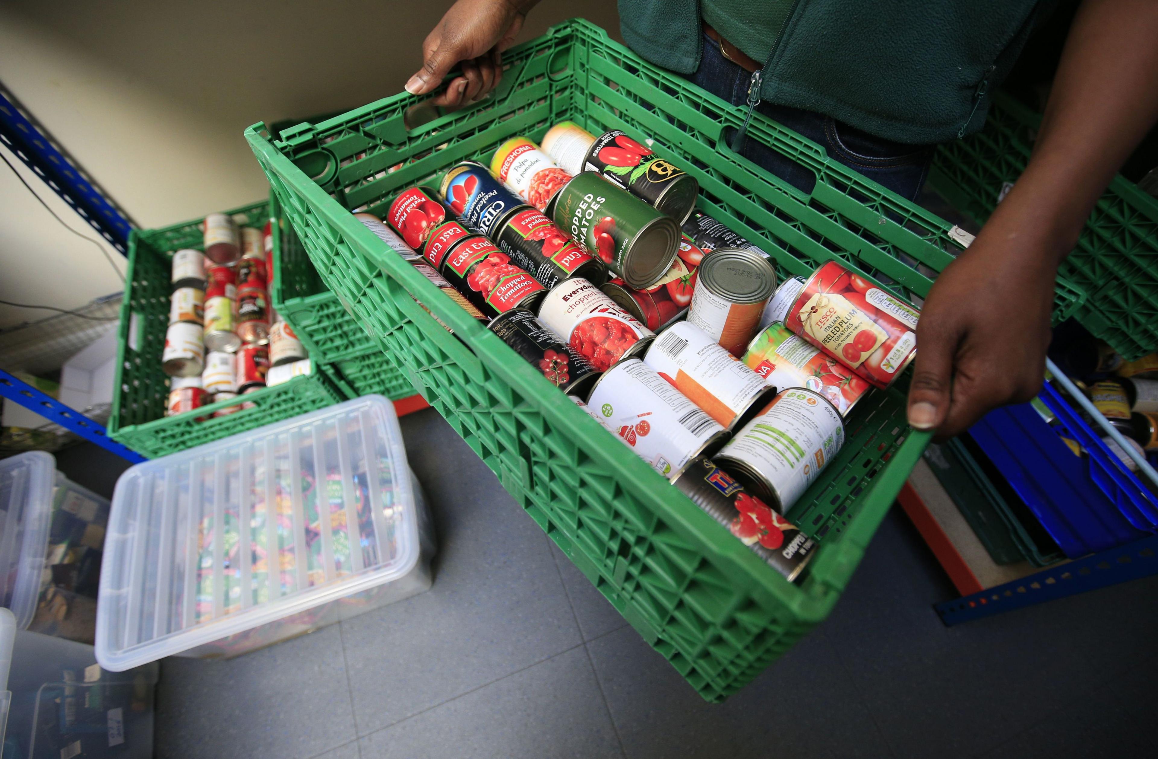 Close up of the hands of a worker at a Trussell Trust food bank carrying a green basket filled with tins of tomato