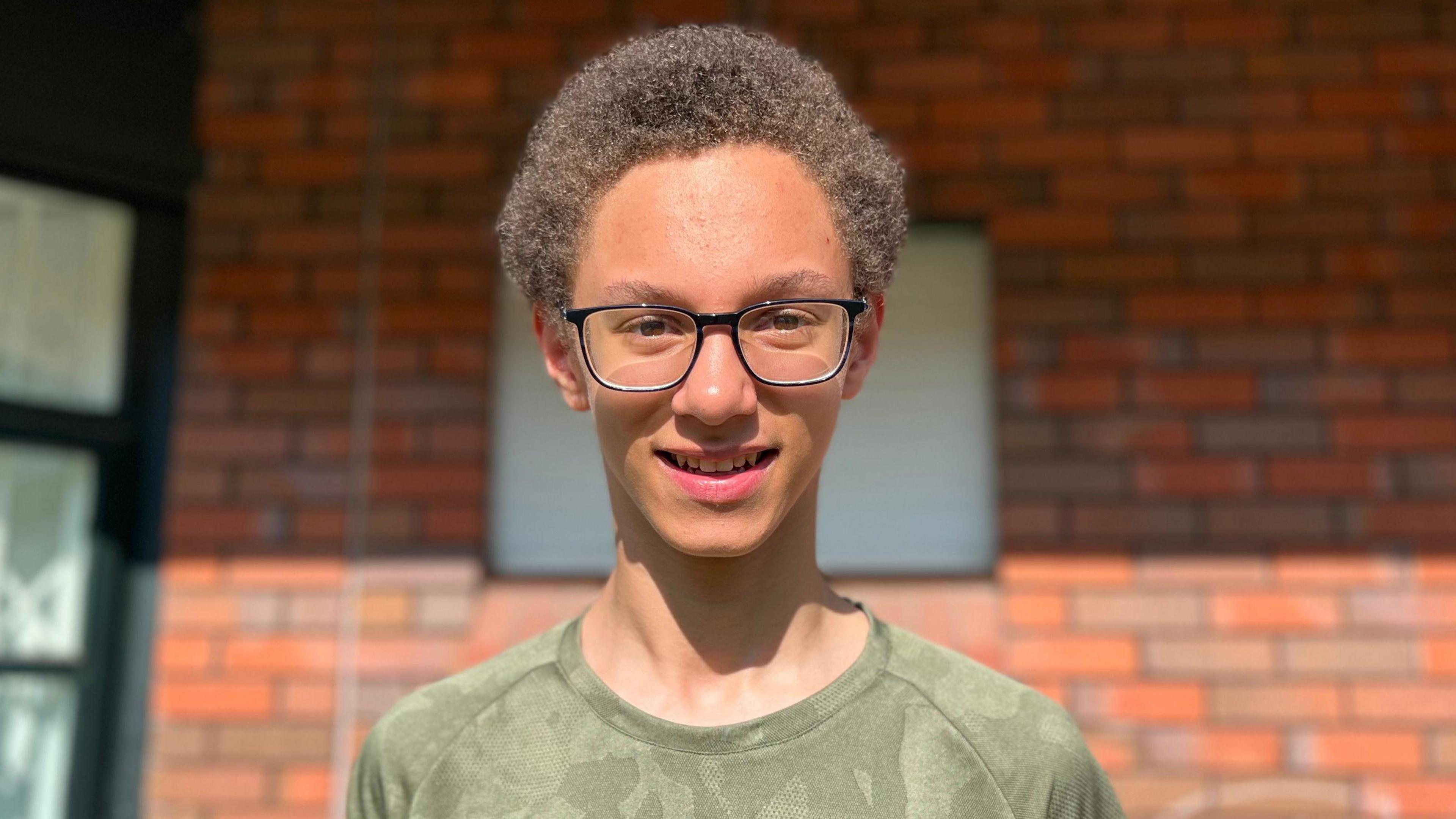 A boy with curly brown hair is wearing glasses and a green camouflage print top. He is standing in front of a brick wall.