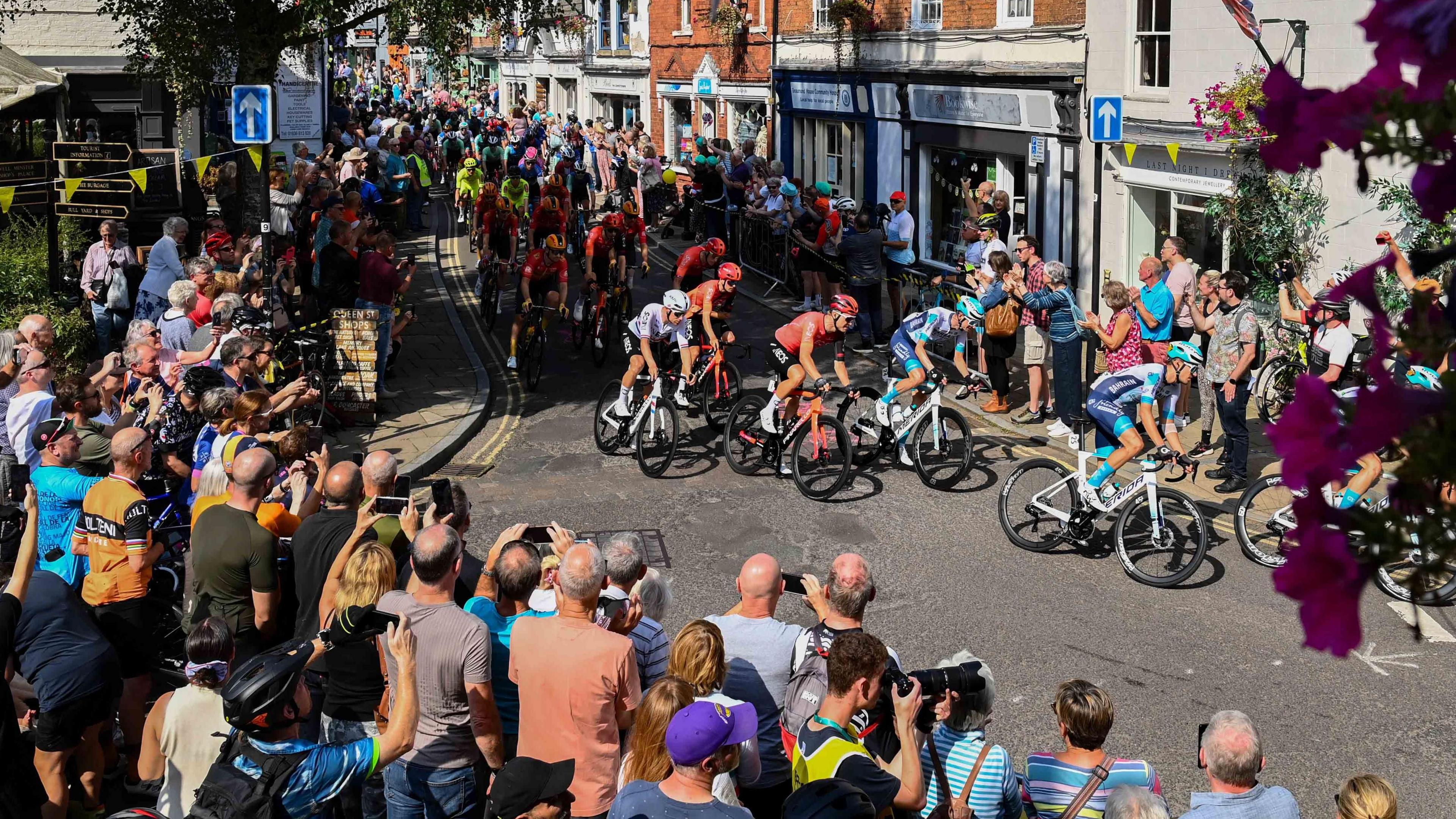 A large group of cyclists riding through a town with crowds cheering them on either side of the street. Some people are taking pictures of the cyclists.
