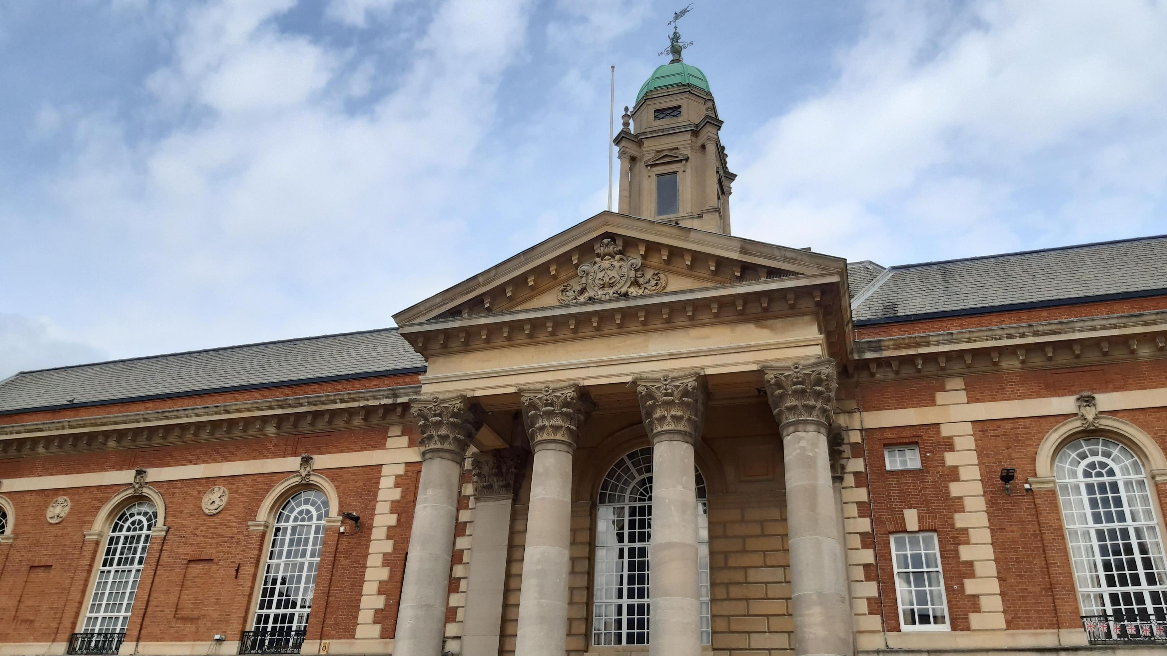 The top of Peterborough town hall. The four pillars outside the entrance are visible. There are blue skies and white whispy clouds.