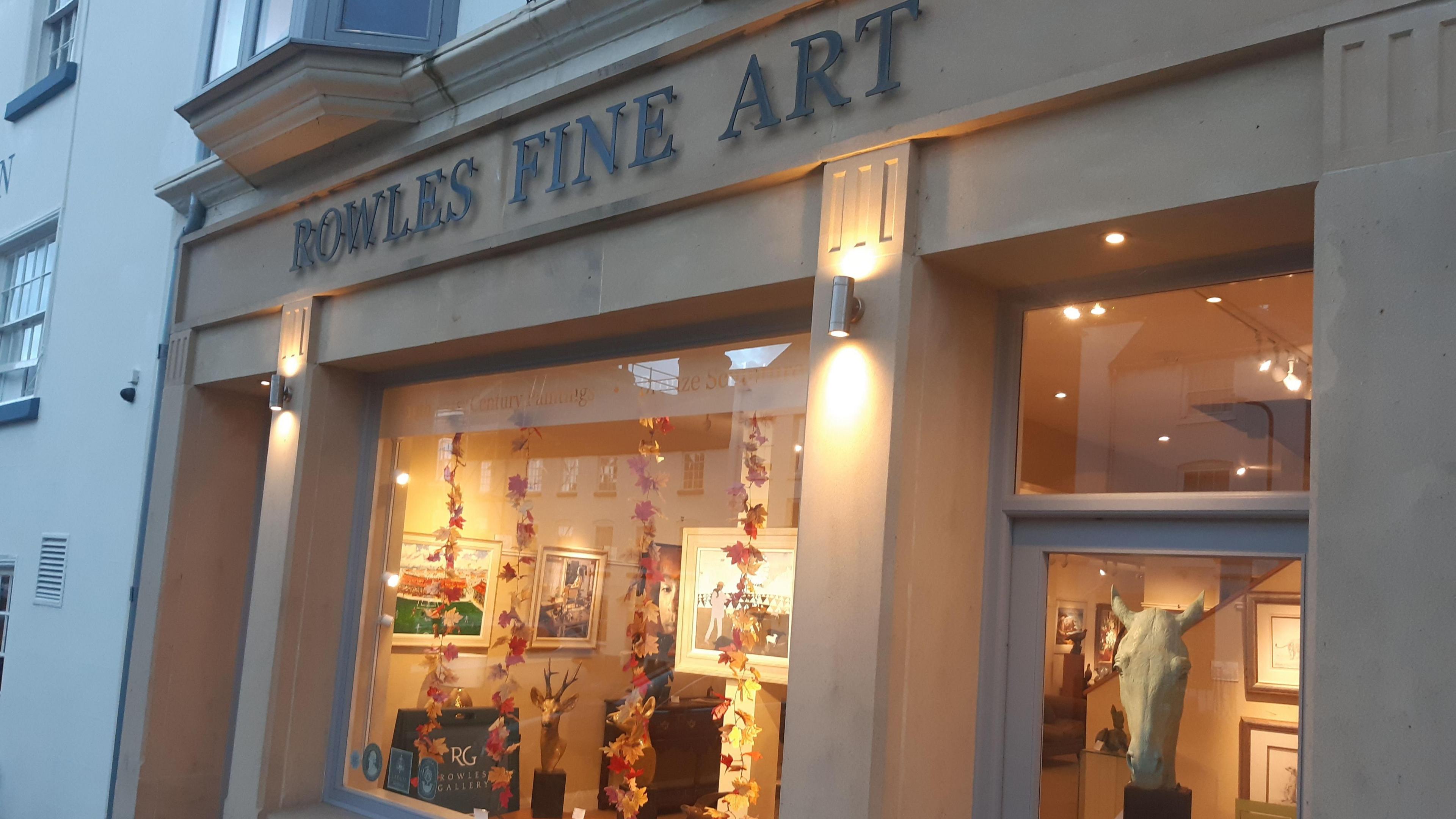 The front of a shop that is gold and has a black sign that reads "Rowles Fine Arts". In the window are orange and yellow autumn leaves dangling in vines, with paintings on the wall inside.
