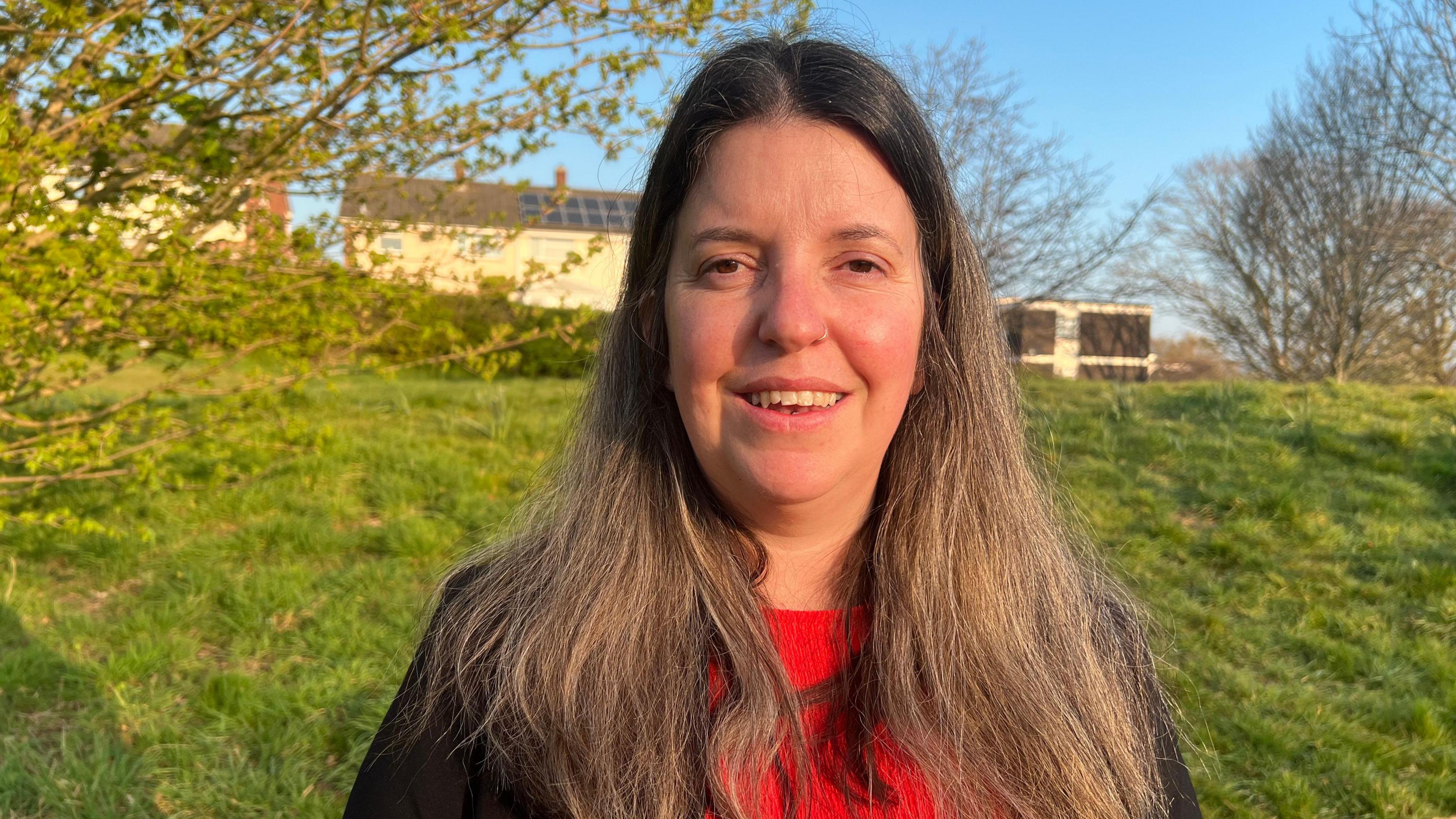 Woman smiles at camera with brown hair and red top. Behind her is a grass bank, some small trees and a house with solar panels on the roof
