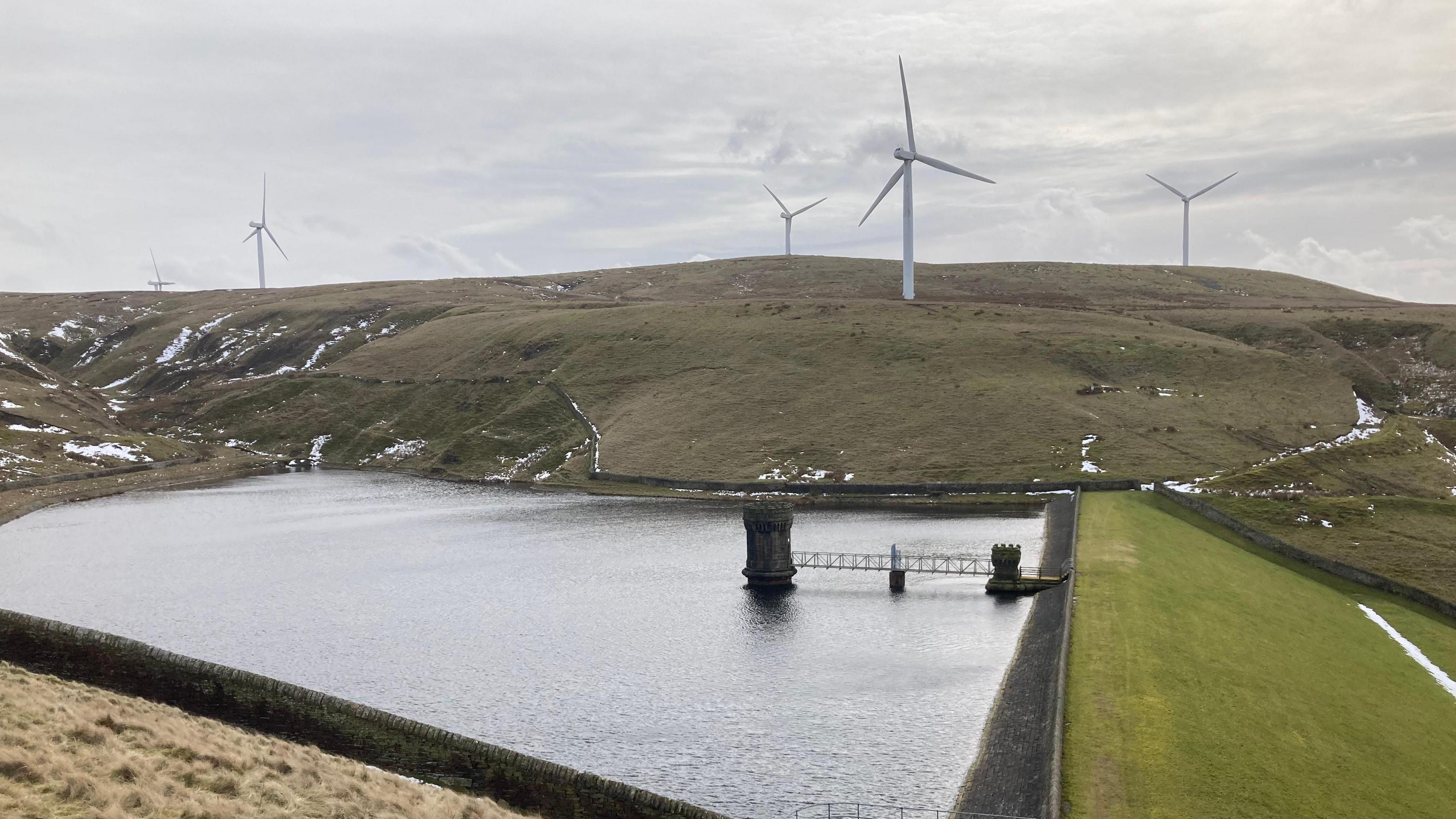 The wind farm of Scout Moor, with turbines seen on headland above a reservoir on an overcast day, with patches of snow seen in the clefts of the hillside.