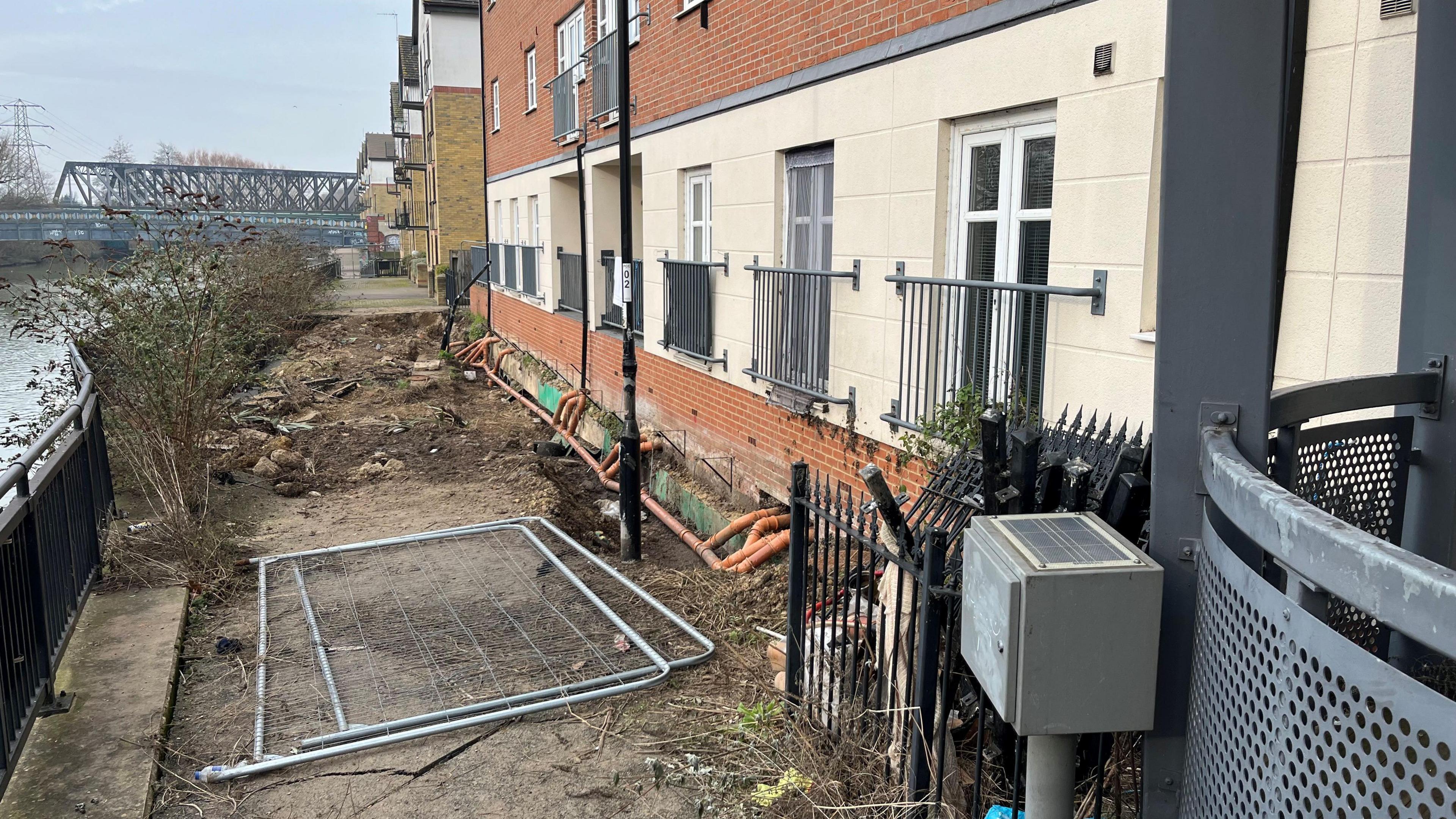 The front of an apartment block near the banks of the River Nene, there is a black wrought iron 1m (3ft) high fence running along the top of the bank that has bulged out in the middle looking like that it might collapse into the water. There are two Heras builders safety fences on the ground and brown sewage pipes can be seen as the ground has been cleared around them. A blue iron railway bridge is visible in the far background on the left.