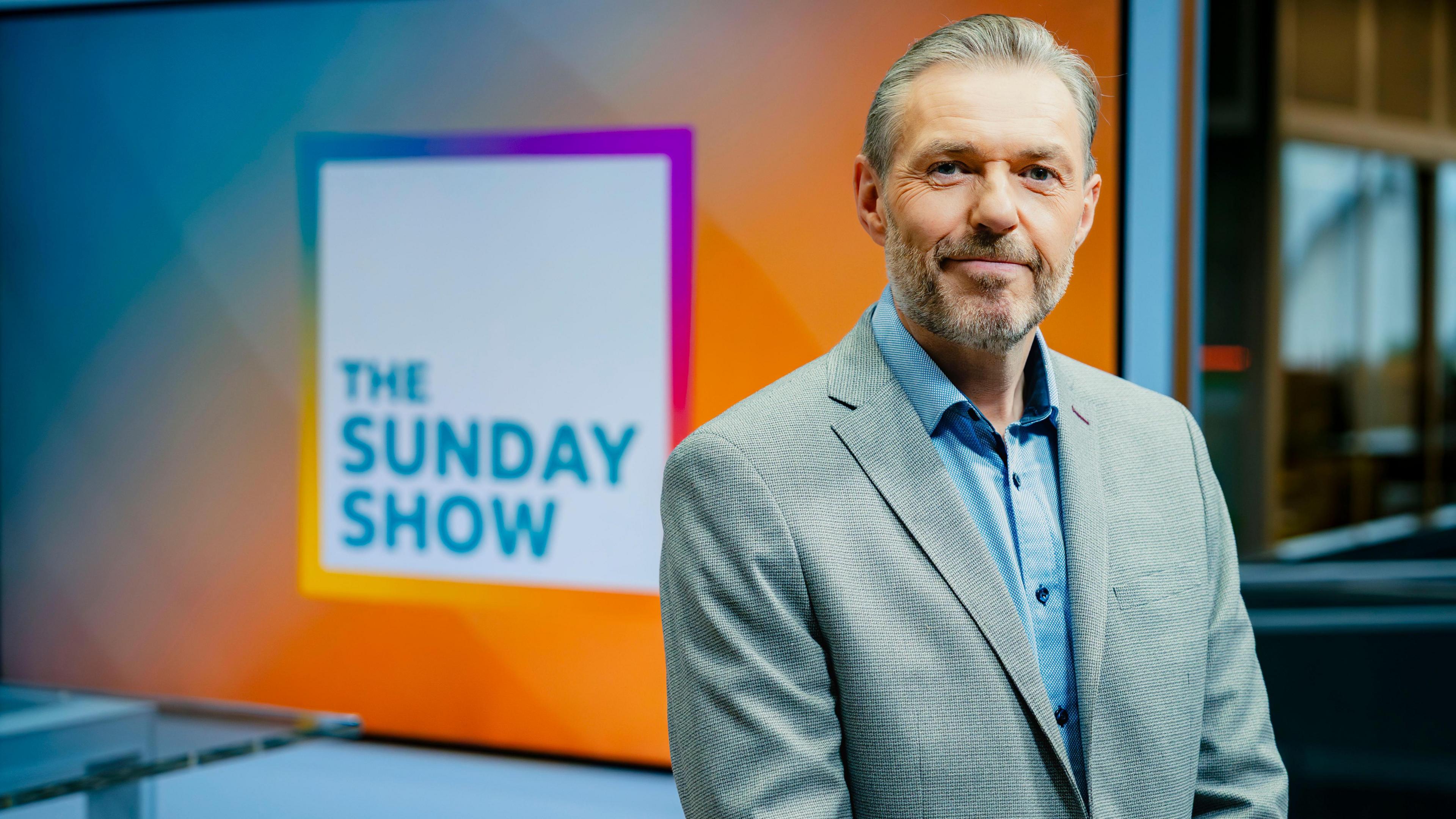 Gary Robertson, a man with grey hair and well-trimmed beard, is smiling while standing on the set of The Sunday Show. He is wearing a grey jacket and blue shirt.