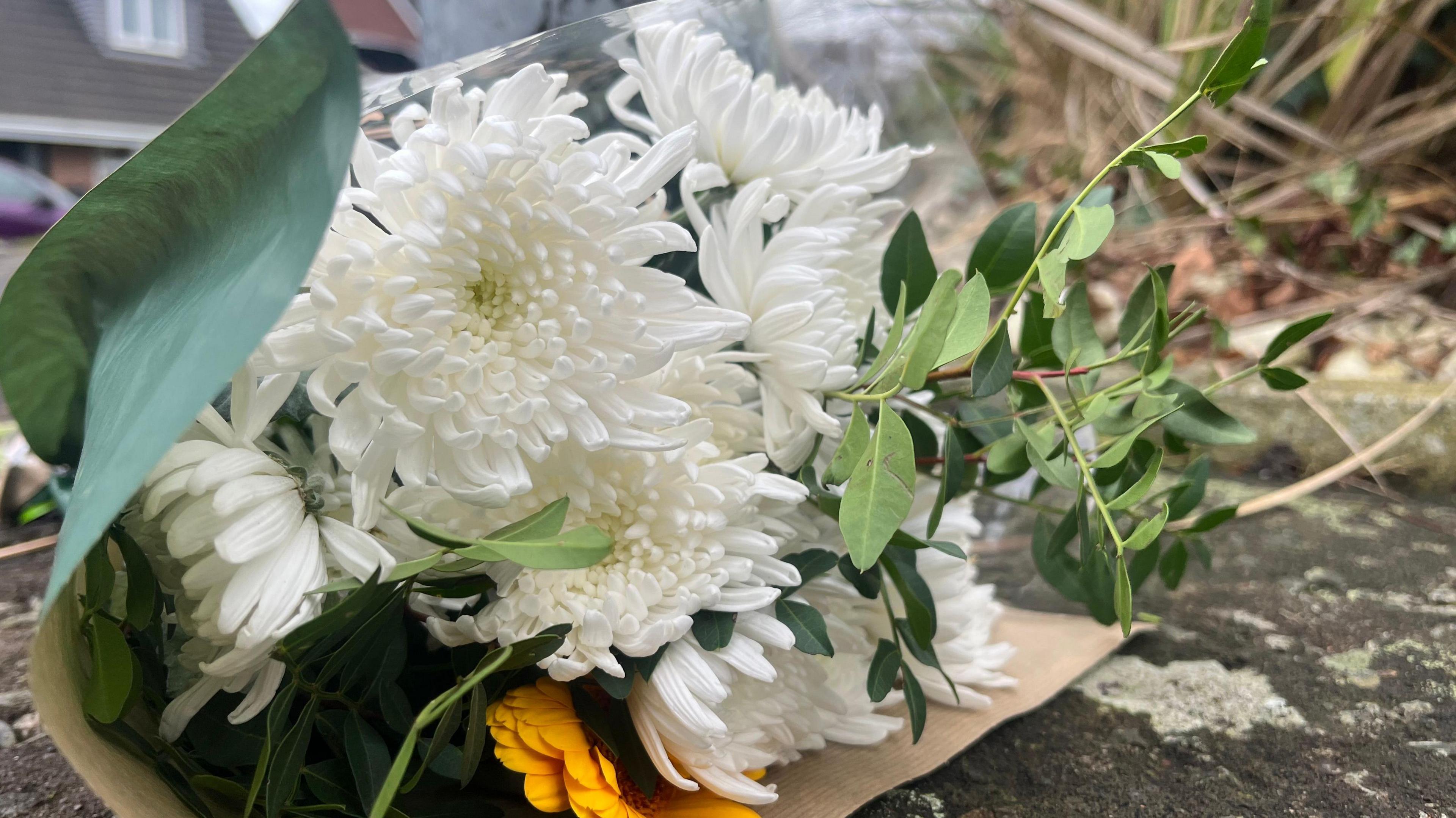 A bouquet of white and yellow flowers with green leaves with green paper wrapping set on a pavement with a house and car in the background.