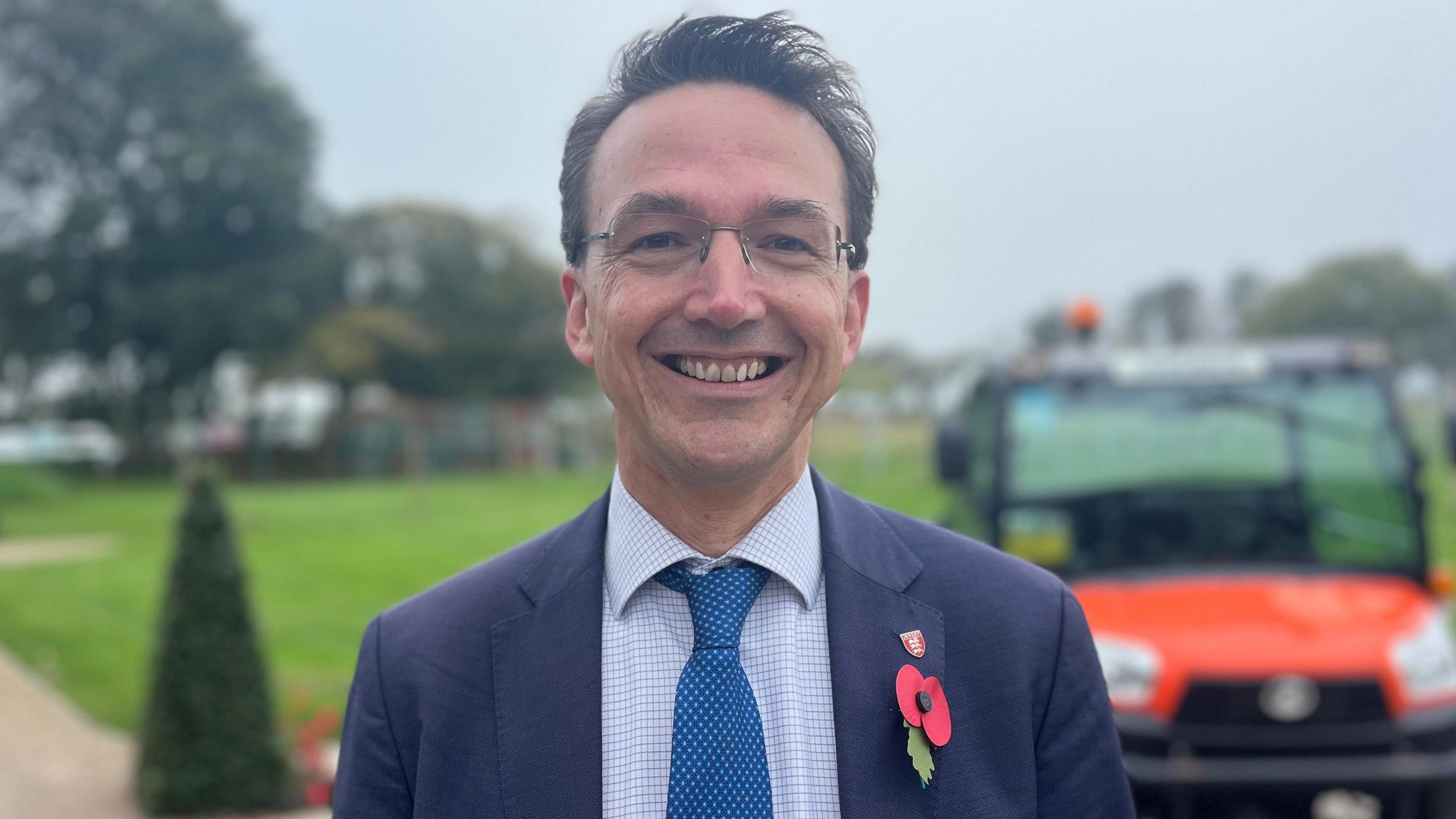 Kirsten Morel is smiling at the camera. He is standing outside with grass, trees and an off-road vehicle blurry behind him. He is wearing a blue jacket, white and blue patterned shirt and a blue tie. He has a Remembrance poppy pinned to his lapel.